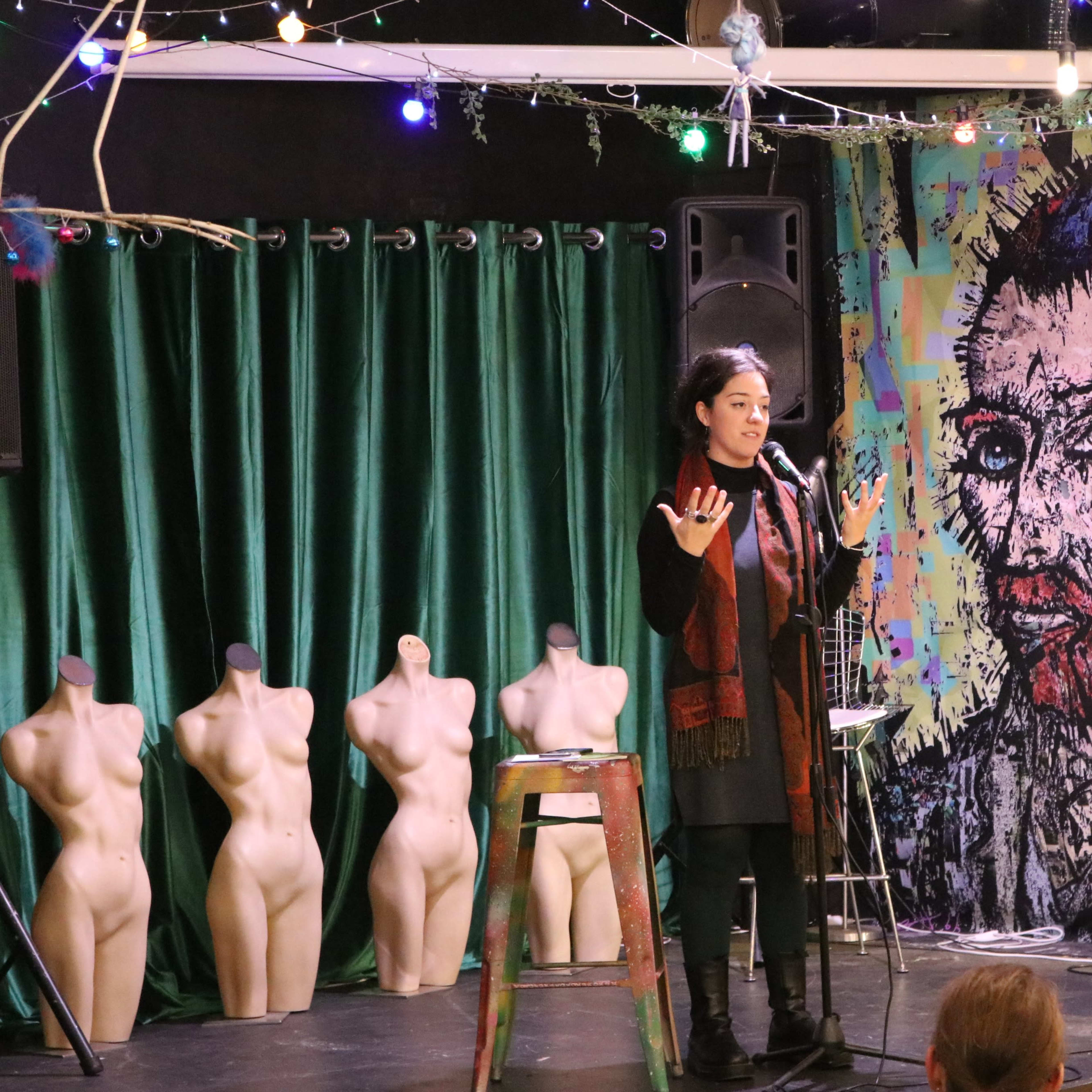 A speaker stands on a stage in front of a colorful mural resembling a famous artist, with green curtains and artistic mannequins in the background.