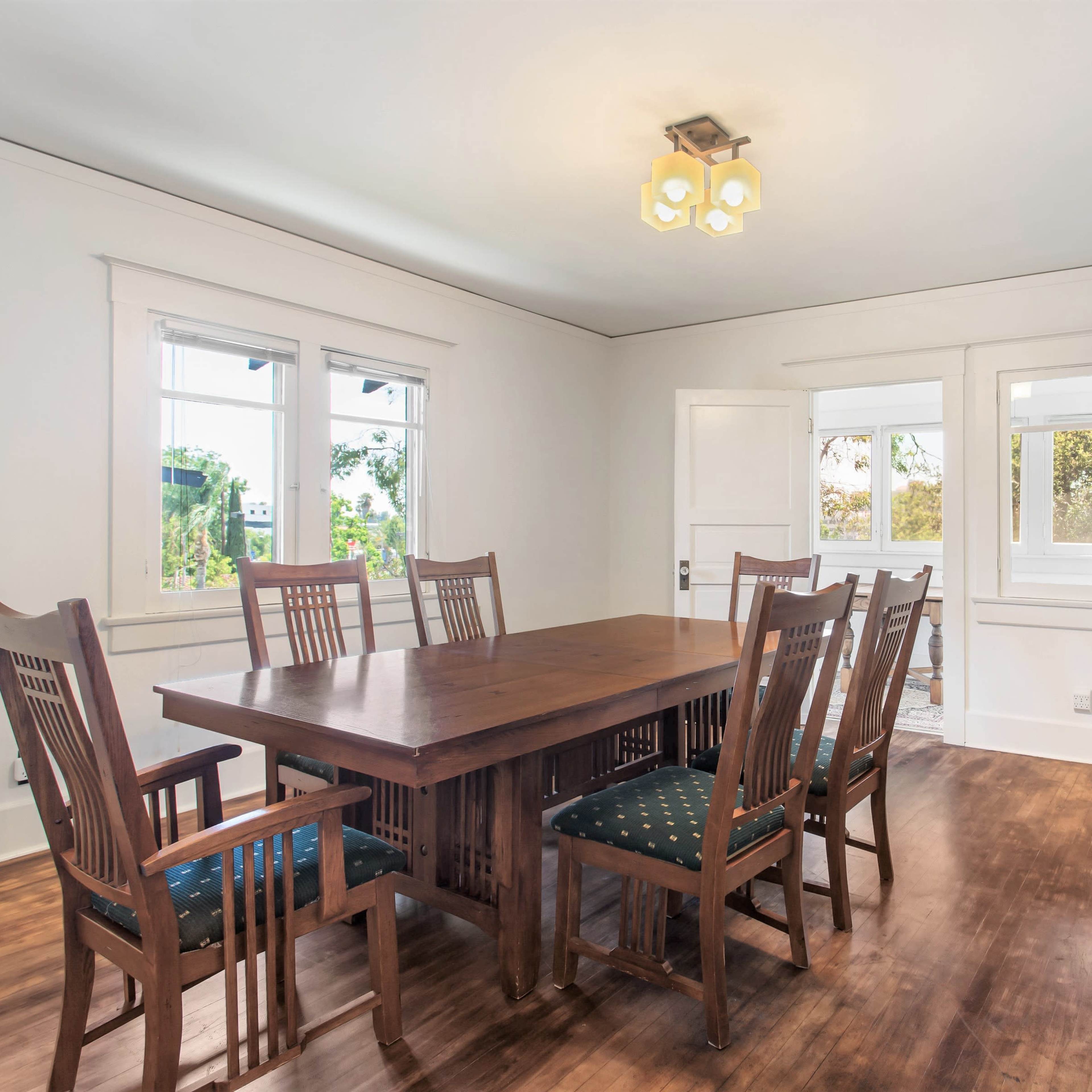 A wooden dining table surrounded by eight chairs is positioned in a well-lit room with large windows and a sideboard.