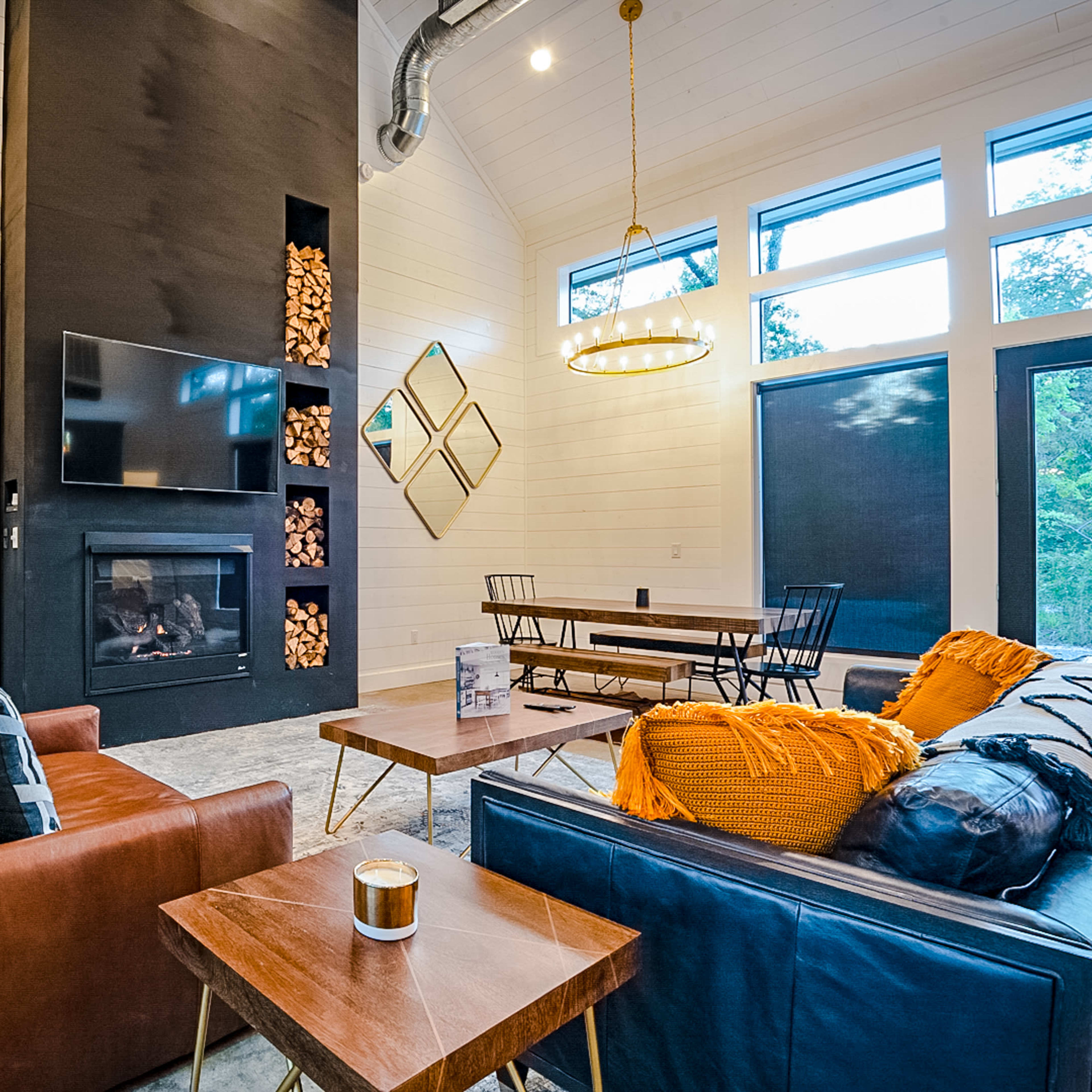 A modern living room with a large black fireplace, a television mounted above it, two couches, a dining table, and large windows allowing natural light.