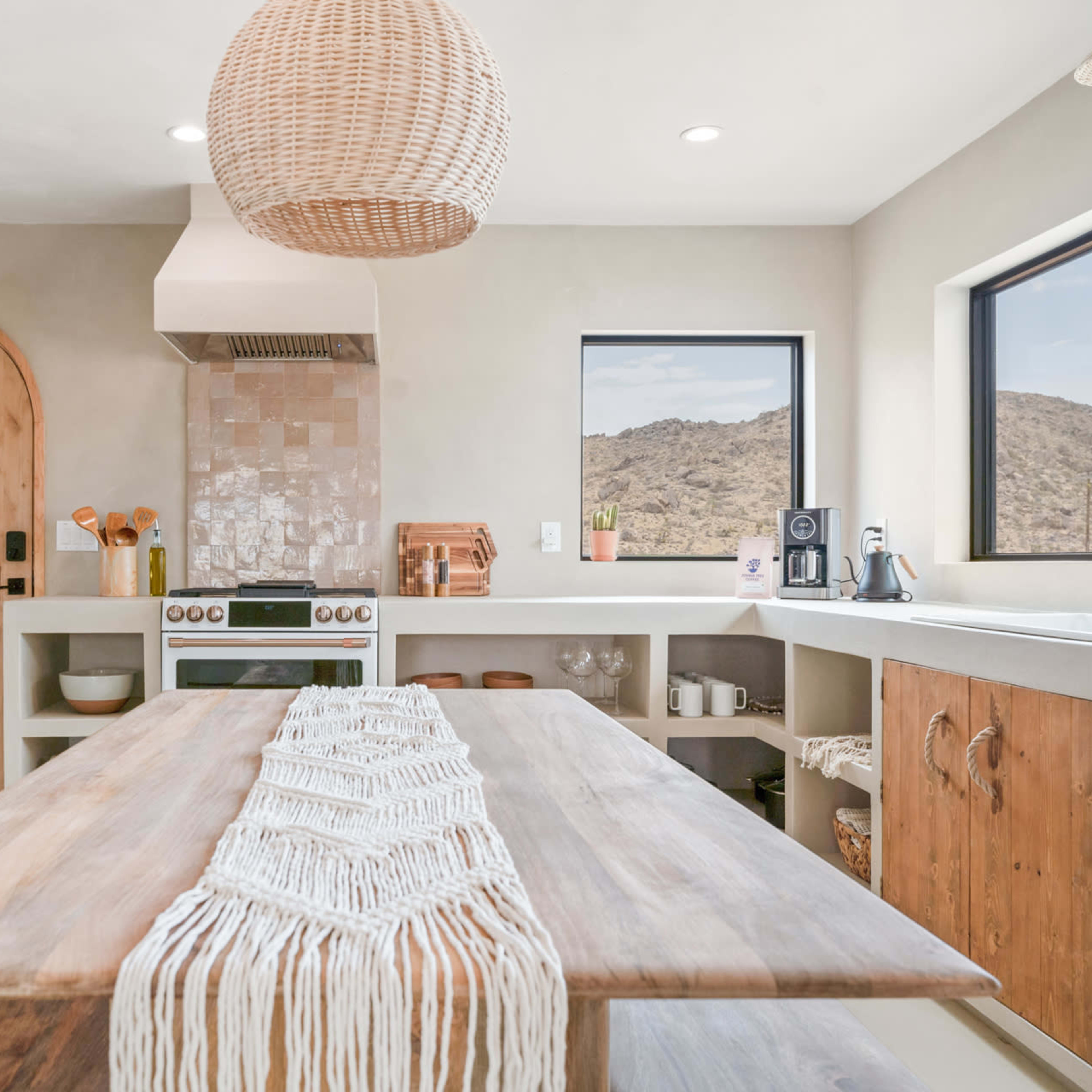 A modern kitchen features a wooden dining table, a round pendant light, and large windows with desert views.