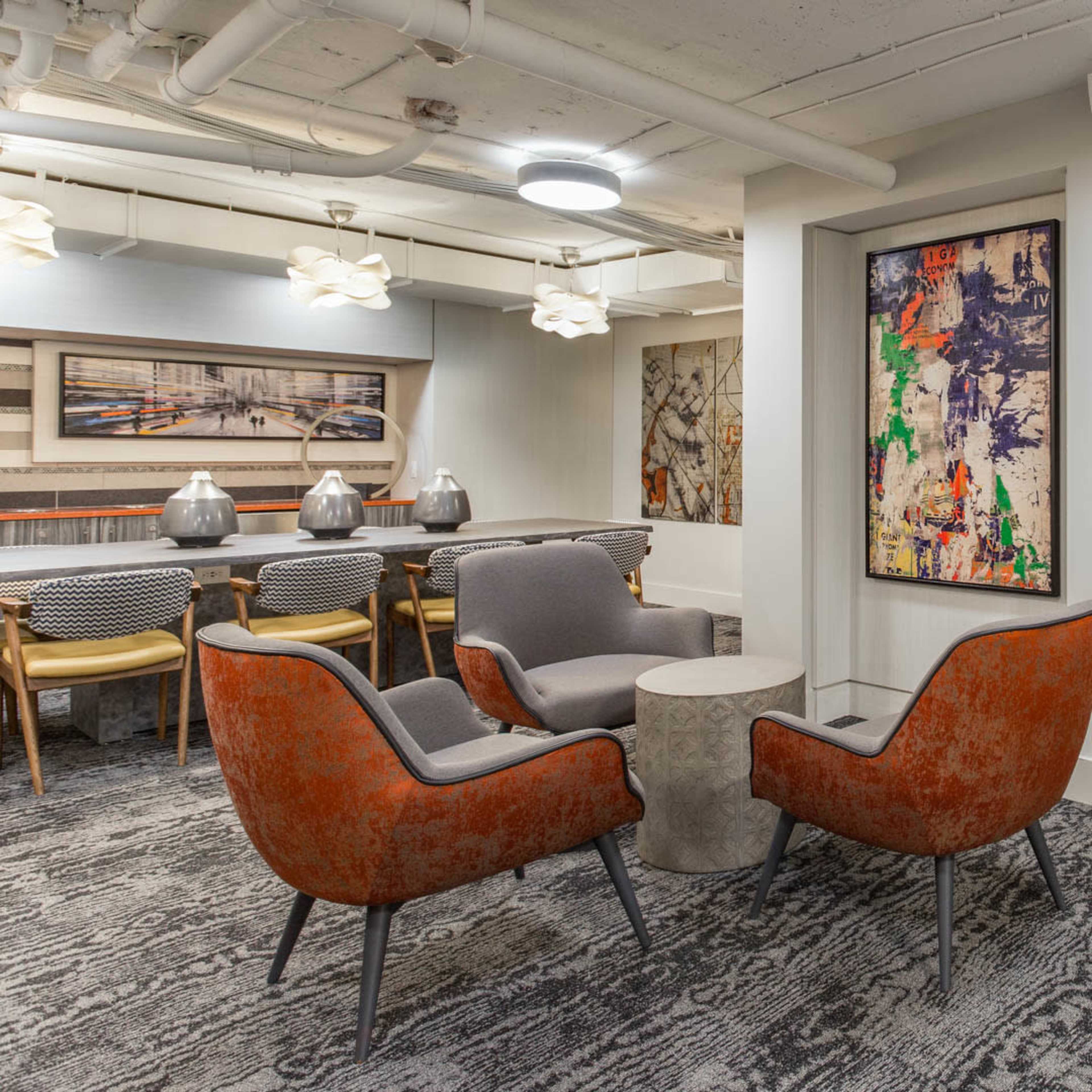 A modern lounge area with a seating arrangement that includes orange accent chairs, a gray armchair, a small round table, and a booth against the wall.