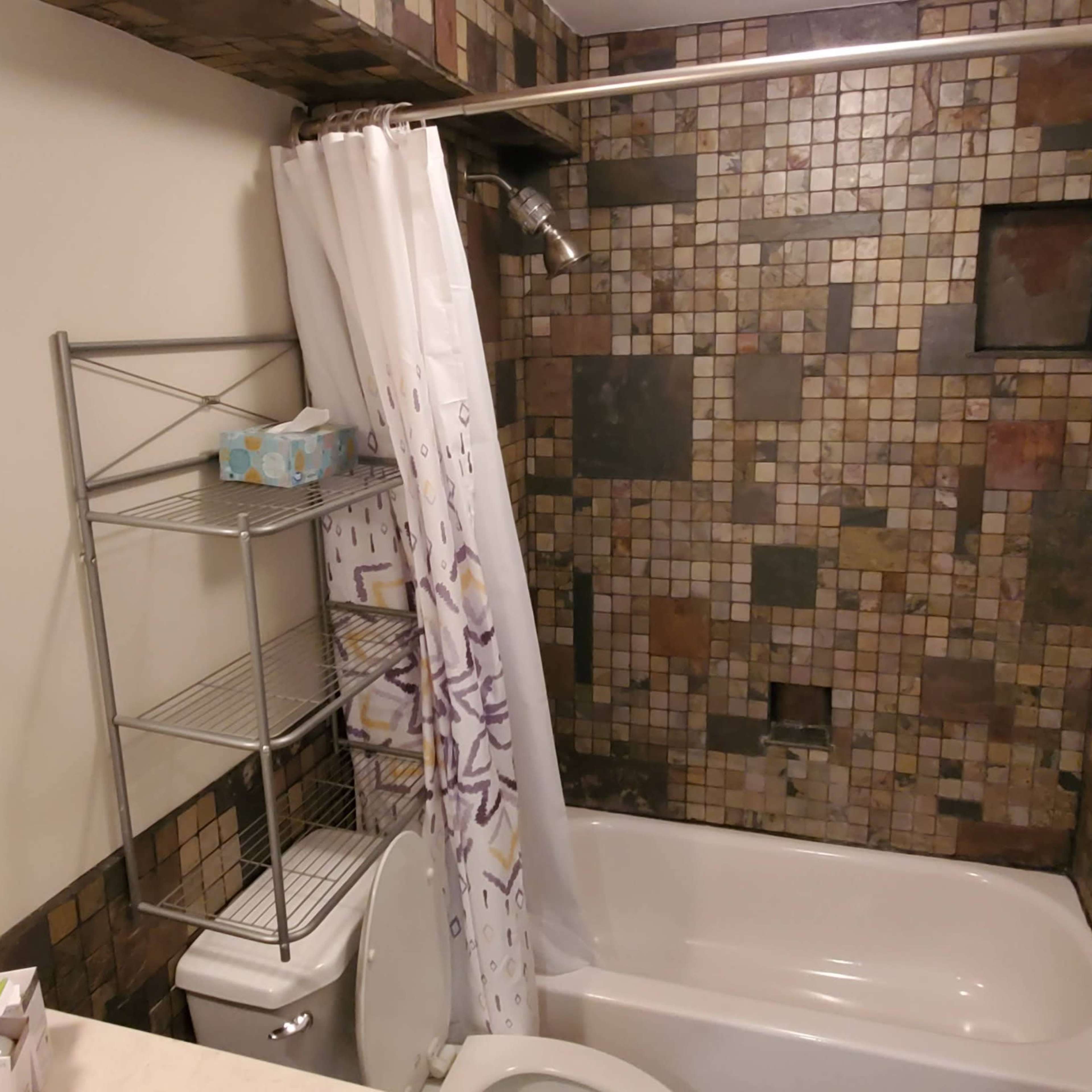 A bathroom with a tiled wall, a bathtub with a shower curtain, a circular mirror, and a metal shelving unit.