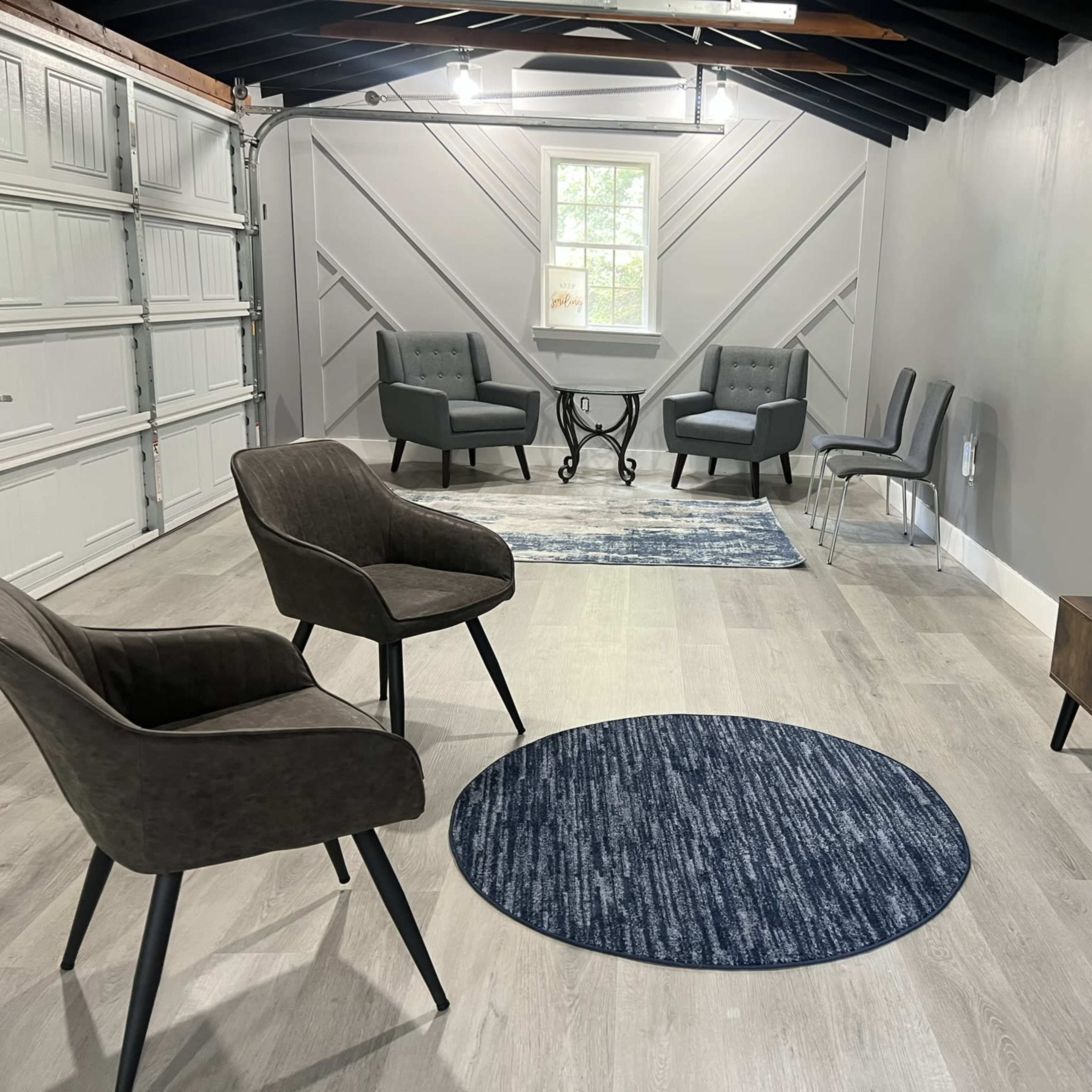 A modern, sparsely furnished room with two types of chairs, a small table, a TV mounted on the wall, and a round rug on the floor.