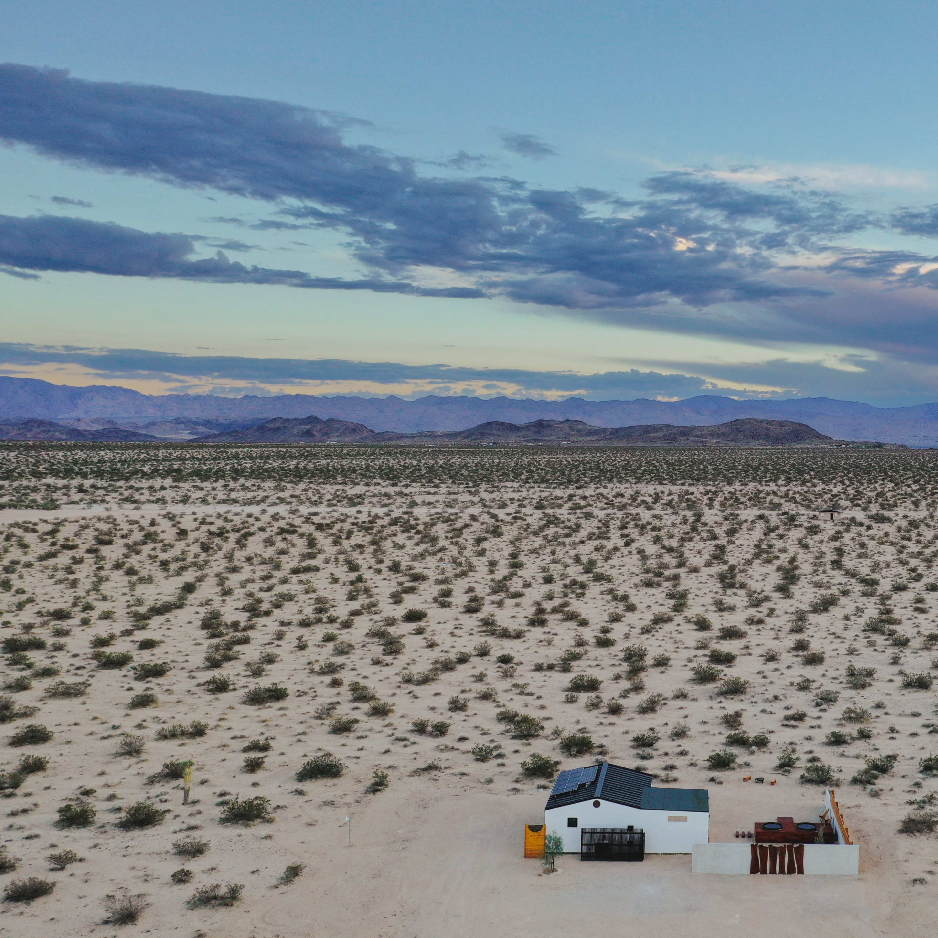 A small white house sits on an expanse of dry desert land surrounded by low vegetation and distant mountains under a cloudy sky.