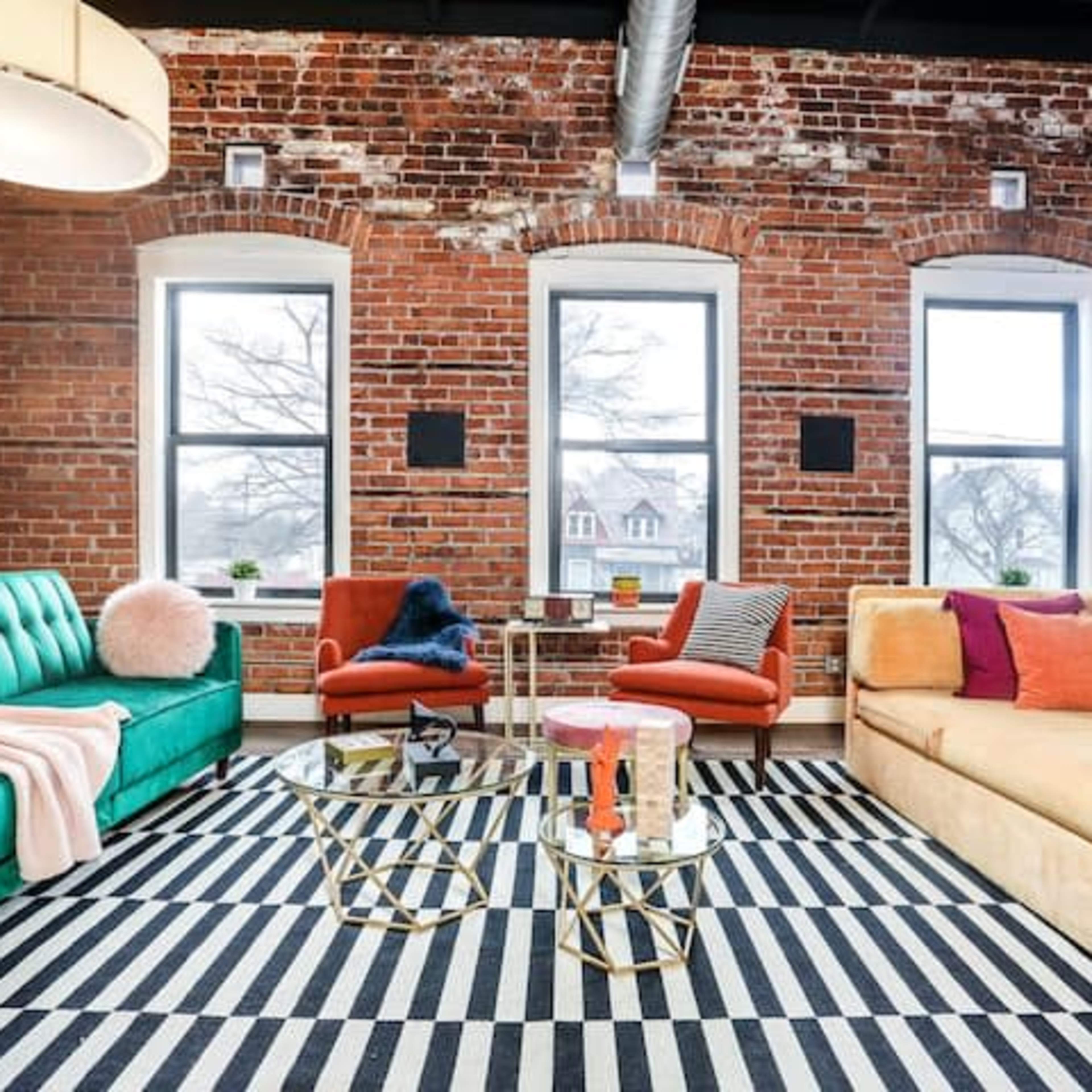 A stylish living room features a green sofa, orange chairs, and a gold and glass coffee table set against a backdrop of exposed brick walls and large windows.