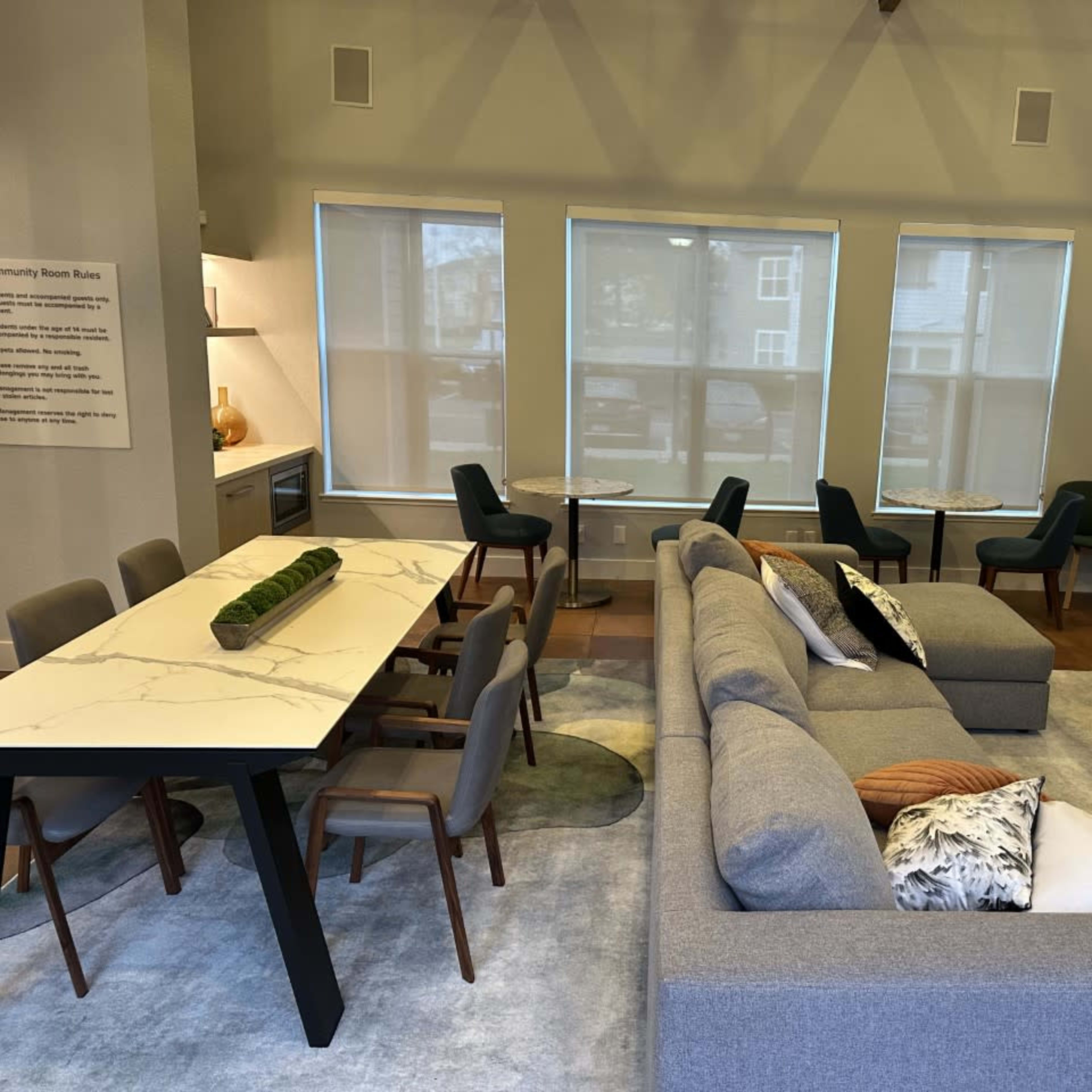 A modern communal lounge featuring a large gray sectional sofa, a marble dining table with chairs, and large windows allowing natural light.