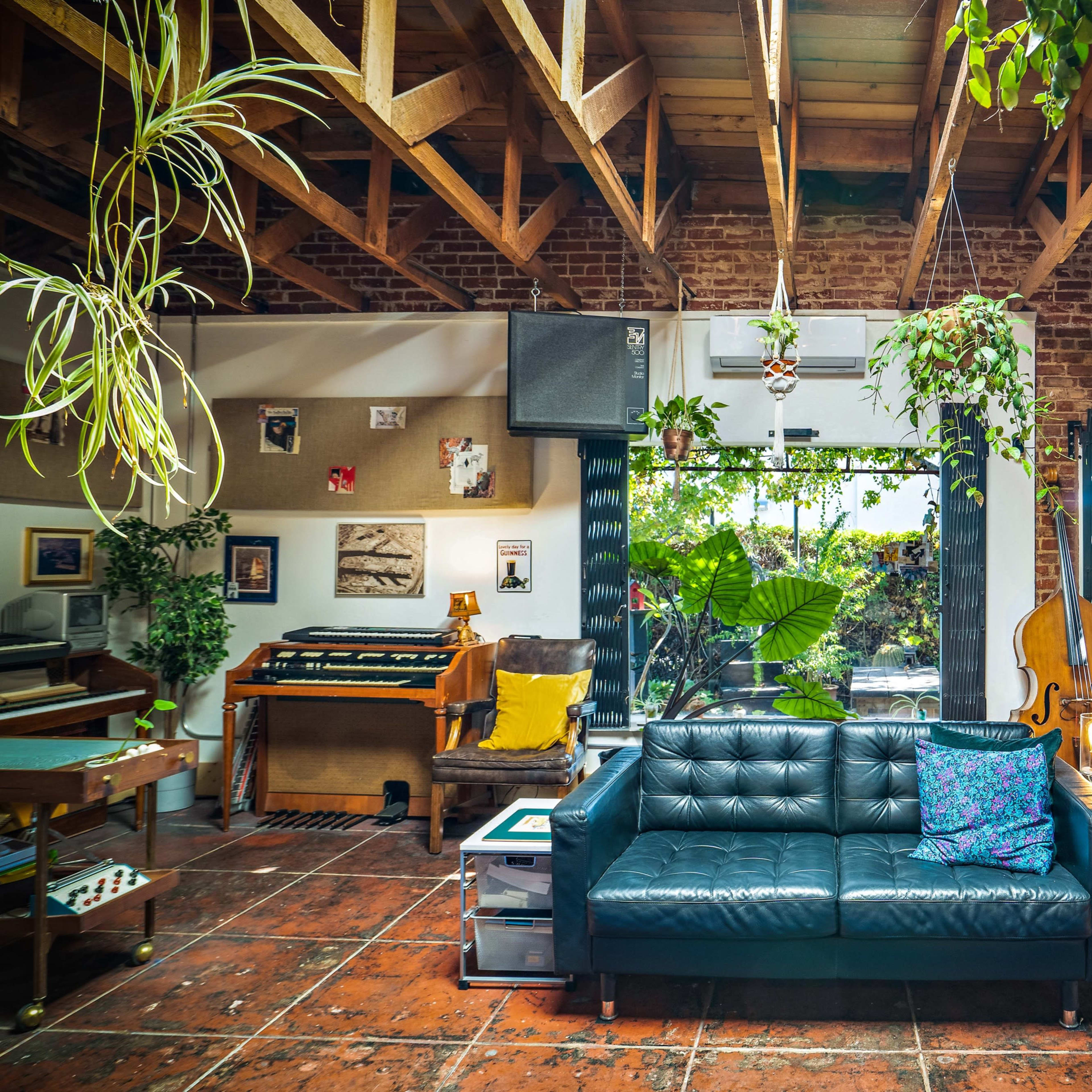 A cozy living room features a black leather couch, musical instruments, and large windows overlooking a green garden.