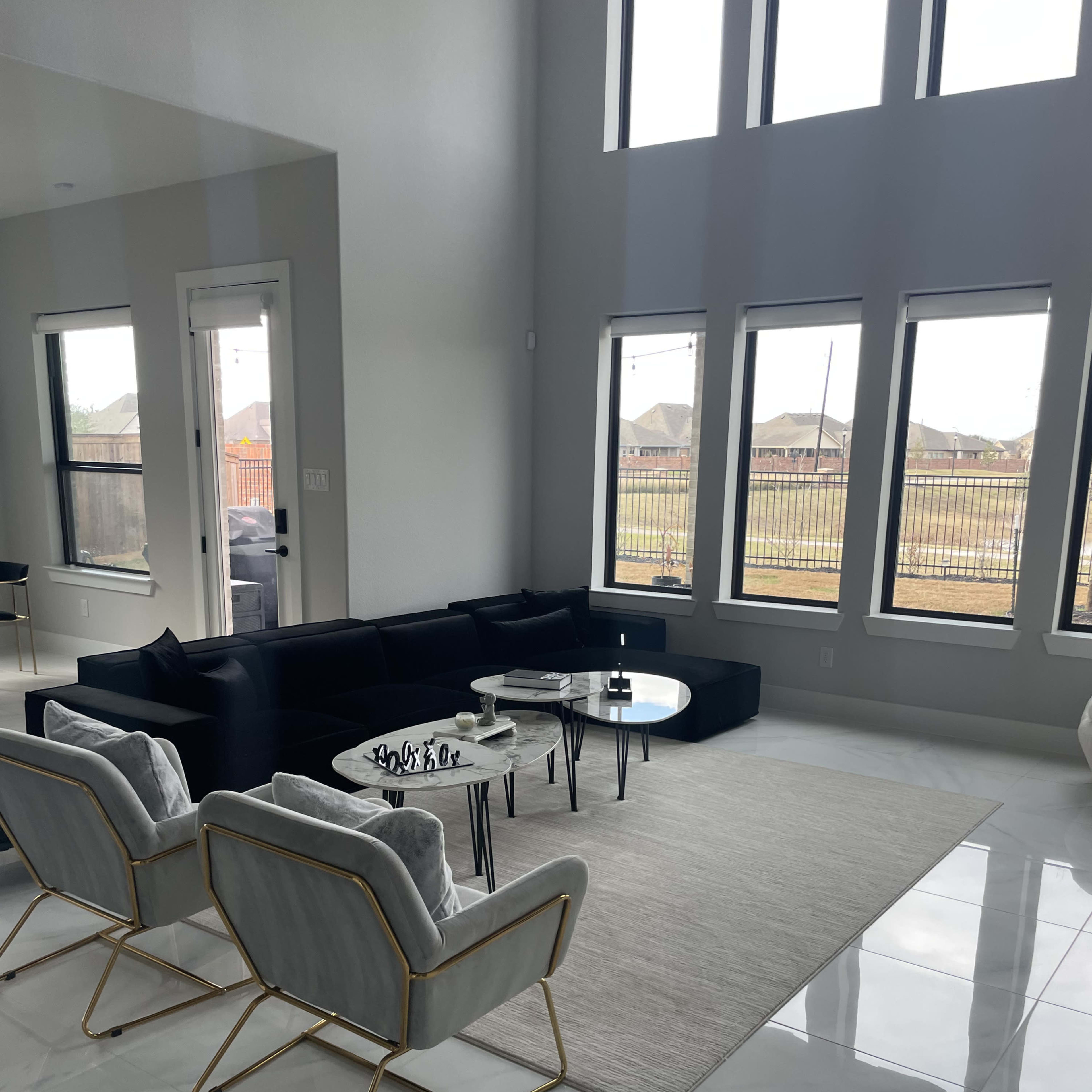 A modern living room with large windows, a black sectional sofa, two light gray chairs, and a round coffee table on a light-colored rug over glossy white tiles.