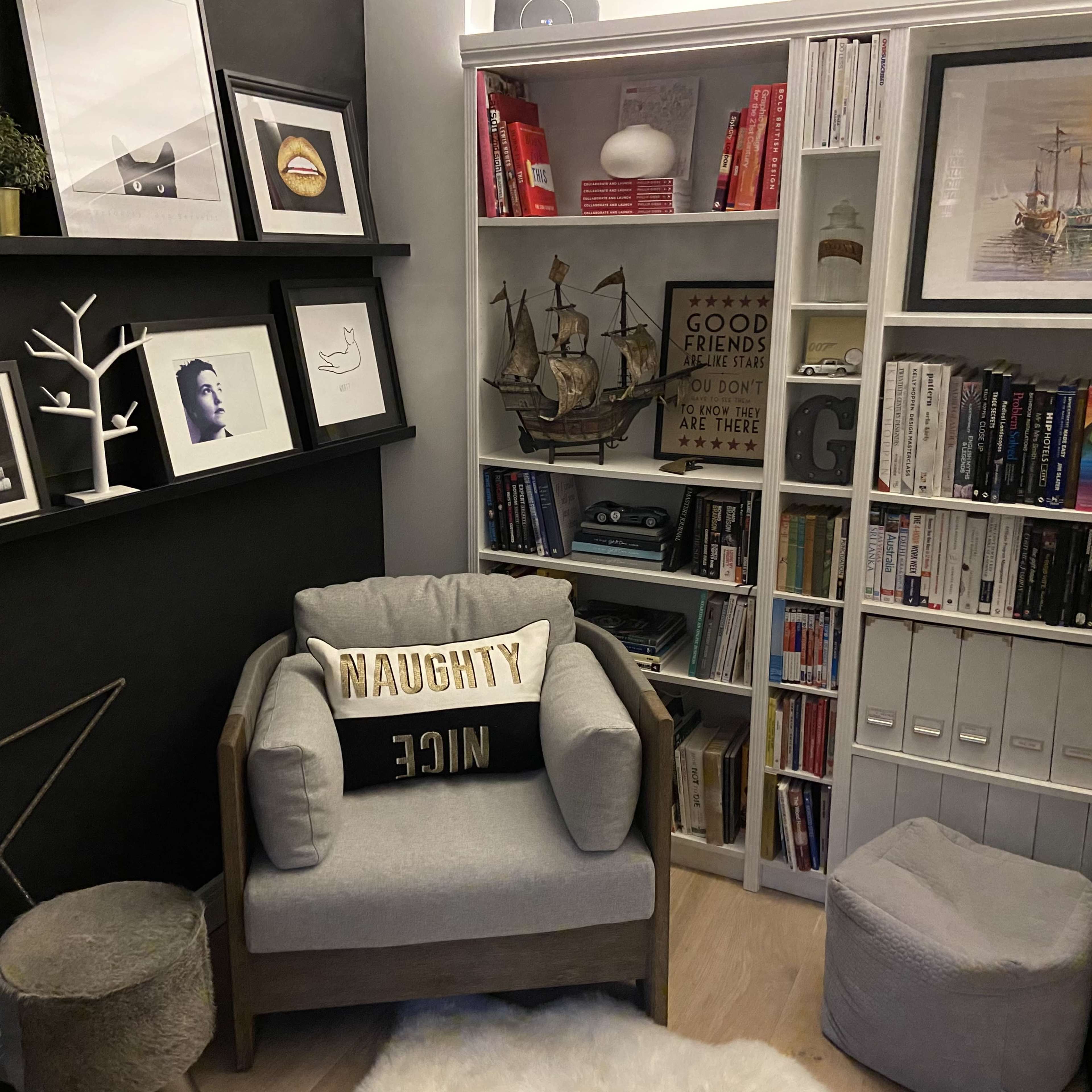 A cozy reading nook featuring a gray armchair with a decorative pillow, a bookshelf filled with books and decor, and framed artworks on the walls.