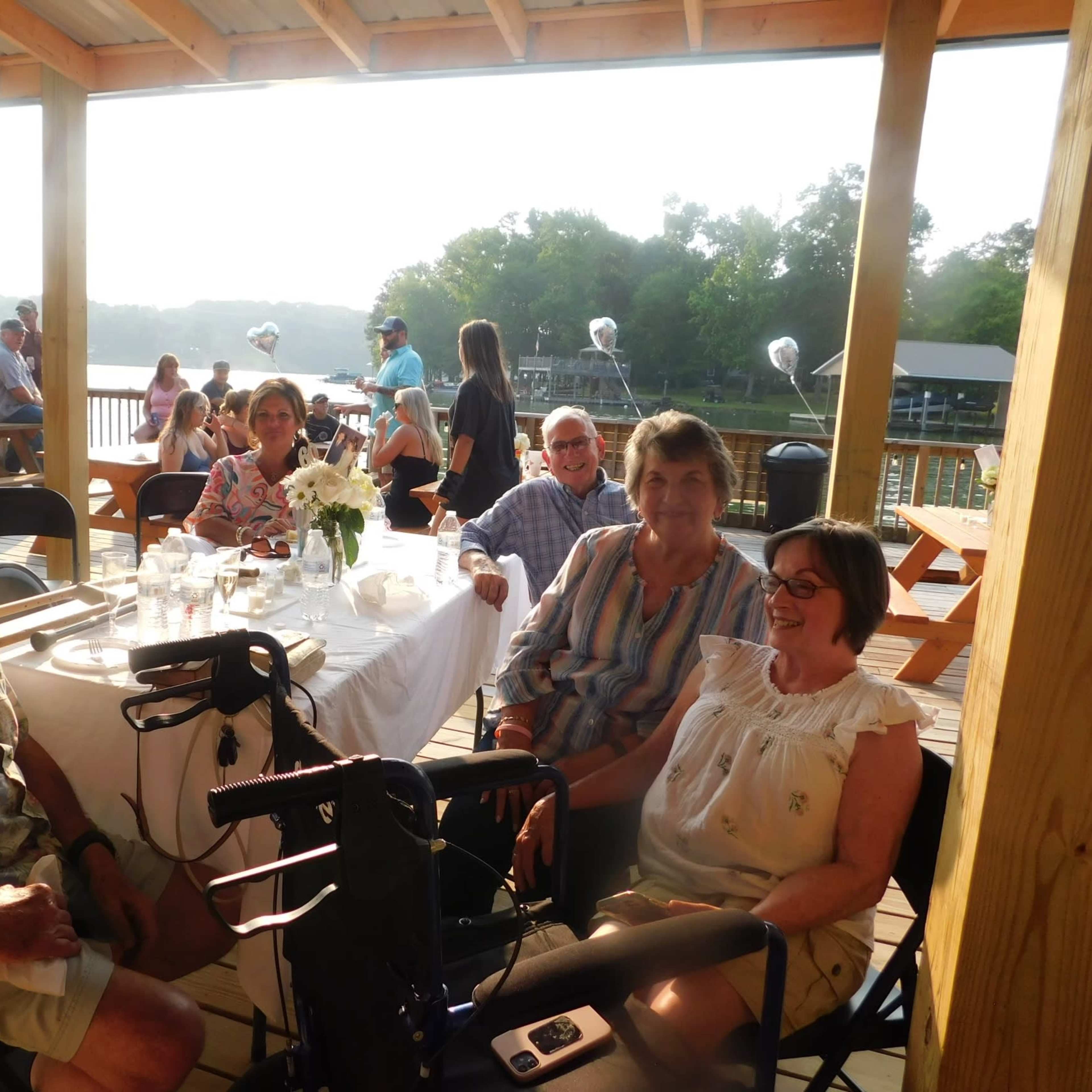 A group of four adults sits at a wooden table on a deck overlooking a body of water, with a festive gathering underway in the background.