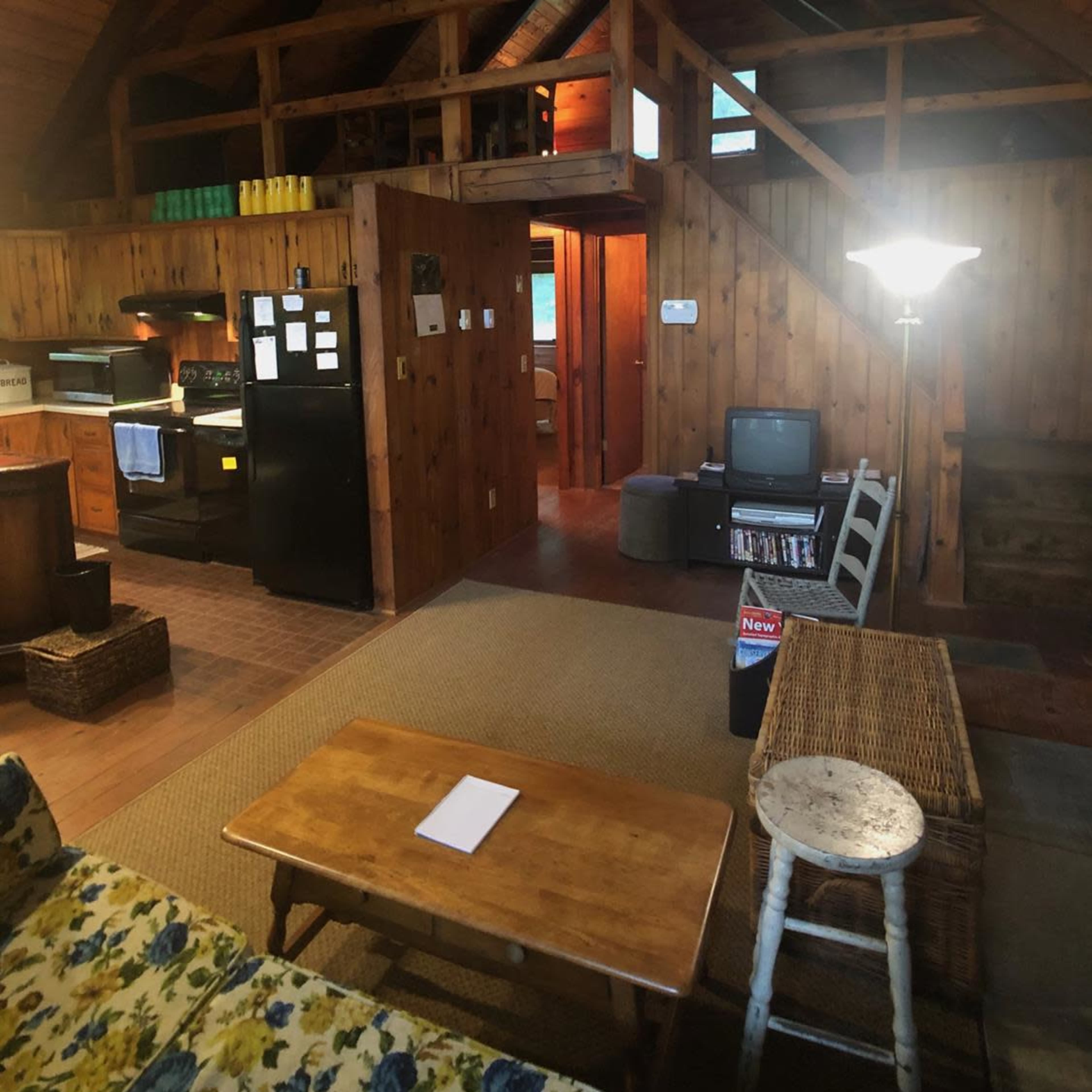 The interior of a wooden cabin features a living area with a couch, coffee table, and a television, alongside a kitchen area and stairs leading to a loft.
