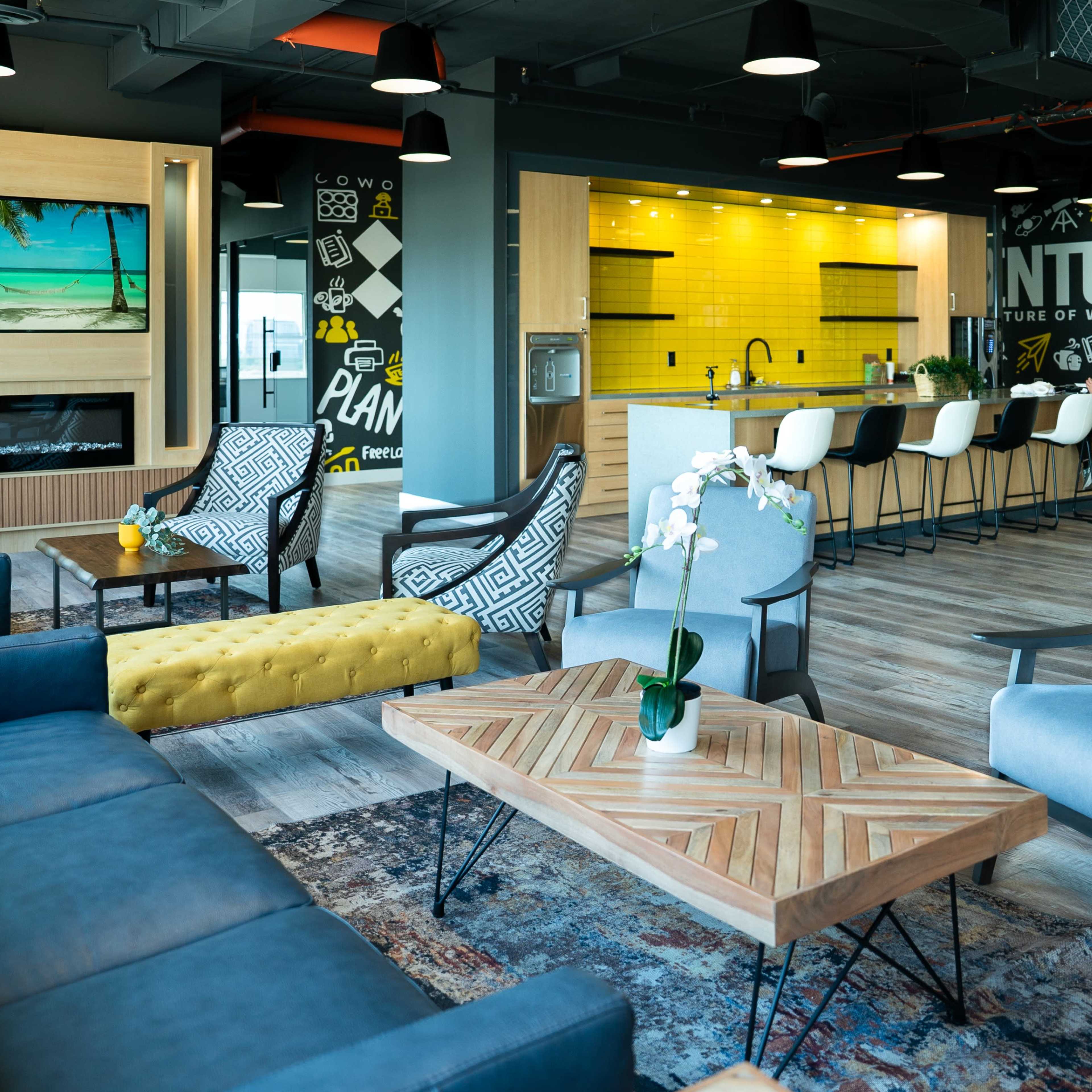 A modern office lounge with comfortable seating, a wooden coffee table, a yellow accent chair, and a kitchen area in the background featuring a mural and a television.