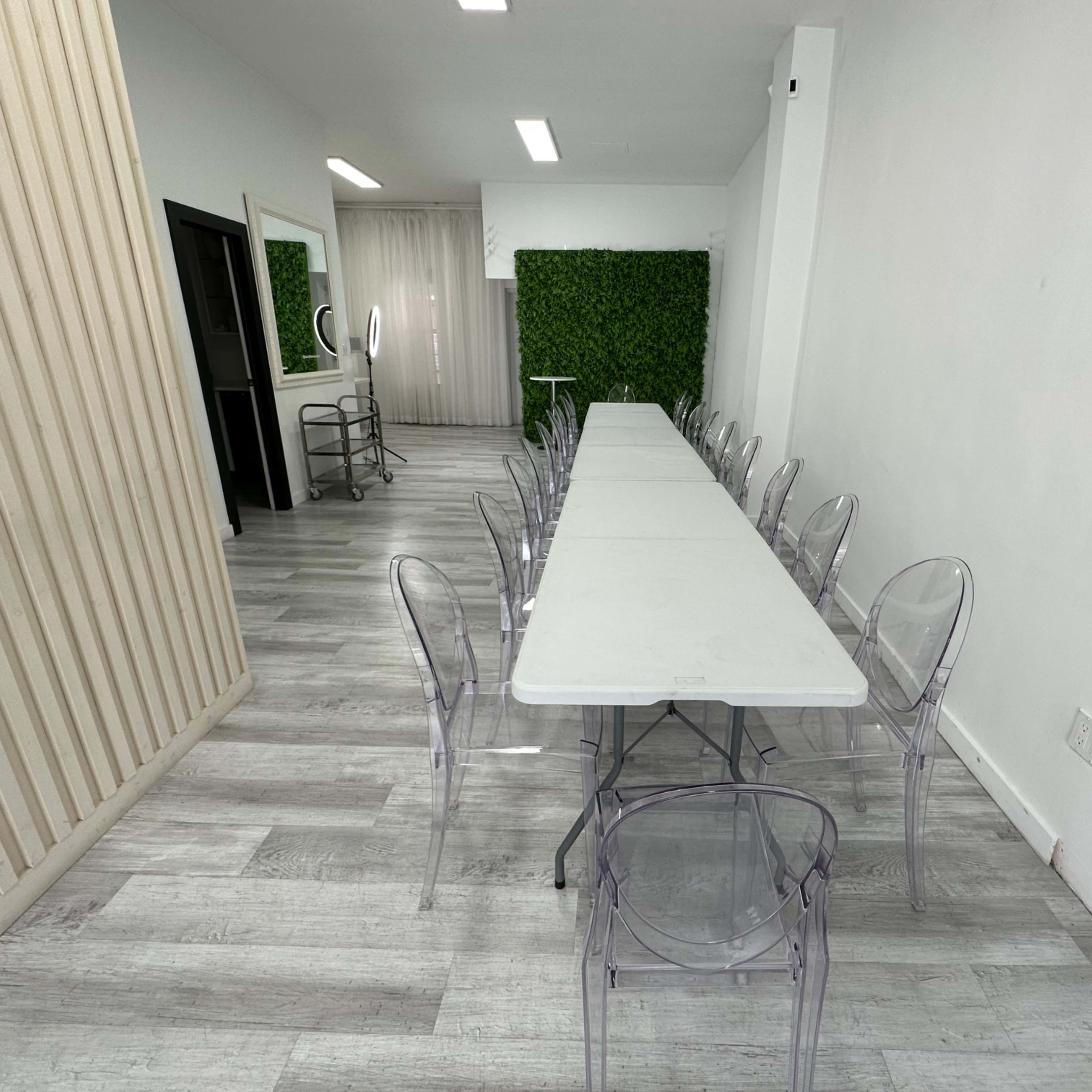A long, white table with clear chairs is set in a bright room featuring a green plant wall and neutral decor.