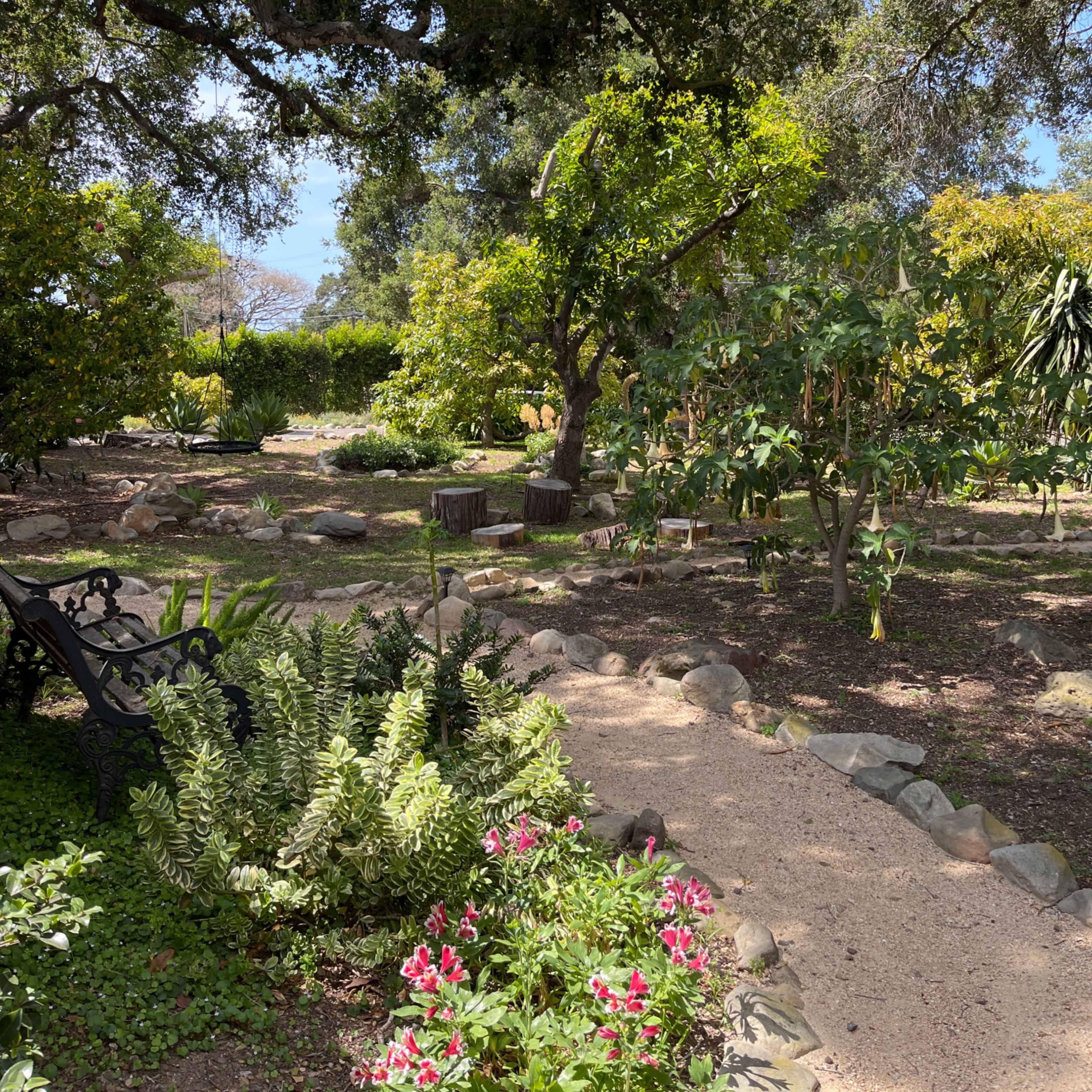 A garden path winds through lush greenery and blooming flowers, with wooden stools nestled under trees.