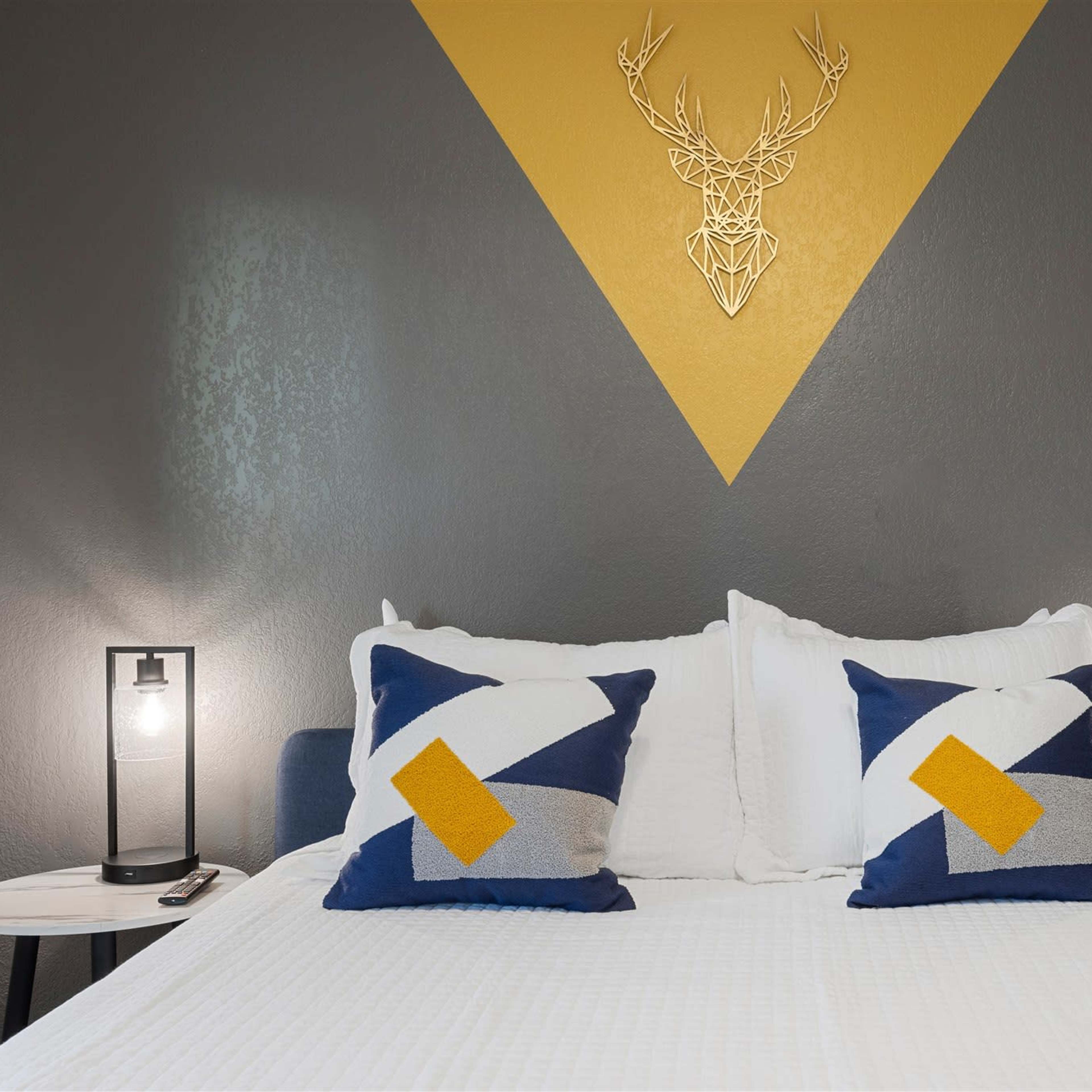 A neatly made bed with two geometric-patterned pillows, flanked by modern lamps, against a dark gray wall featuring a yellow triangular design with a deer head decoration.