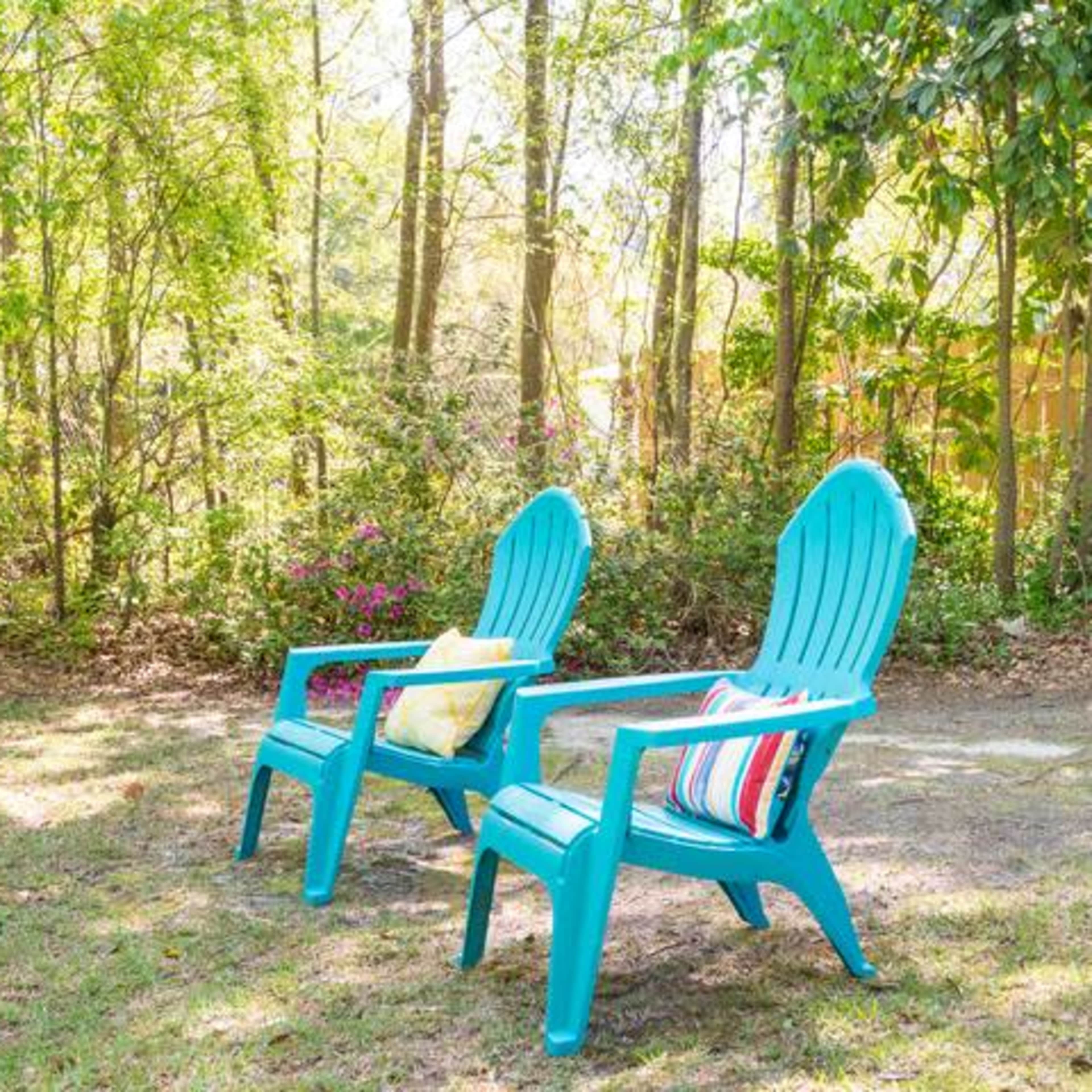 Two teal Adirondack chairs with colorful cushions are situated on a grassy area surrounded by trees and shrubs.