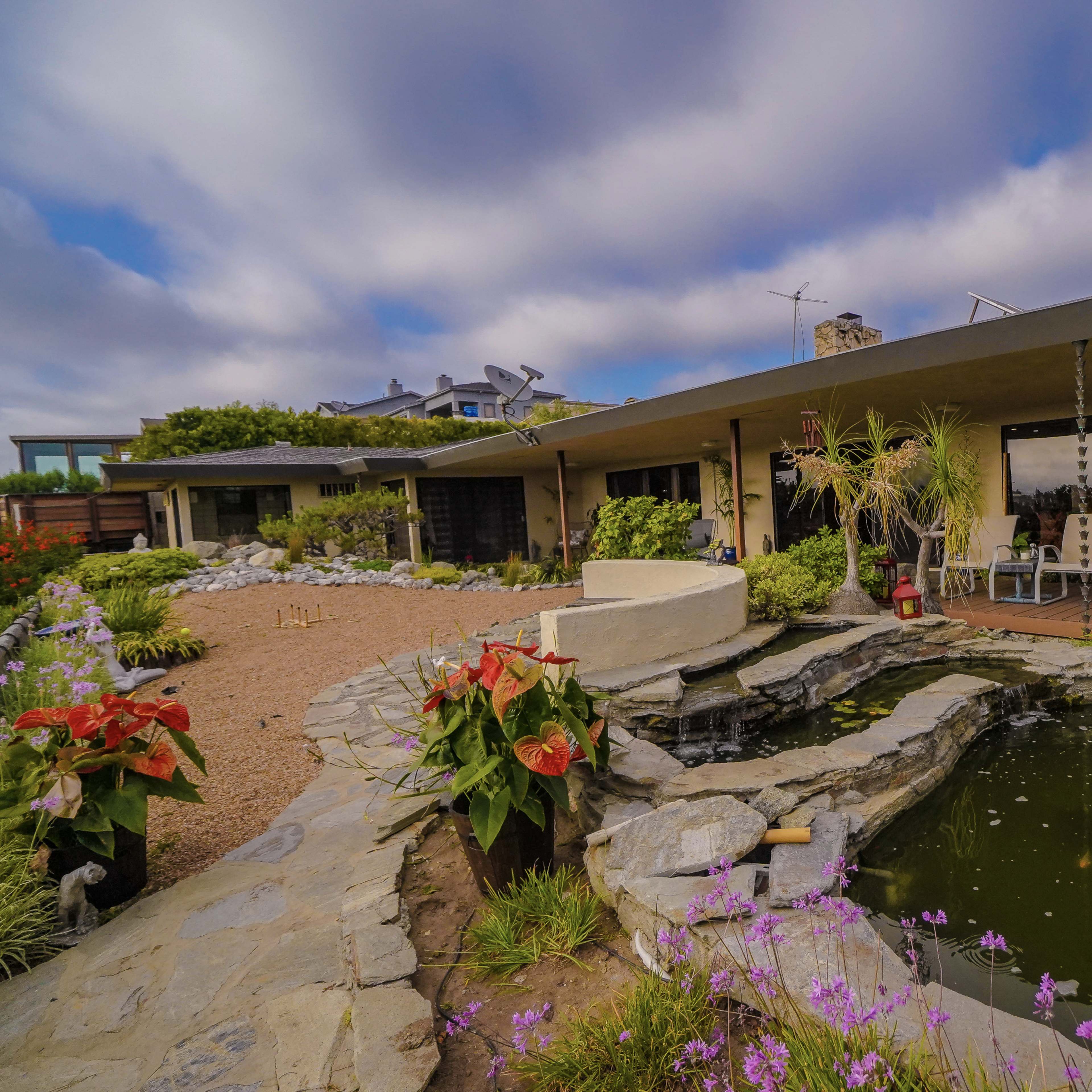 A modern house with a pond and surrounding gardens sits under a cloudy sky.