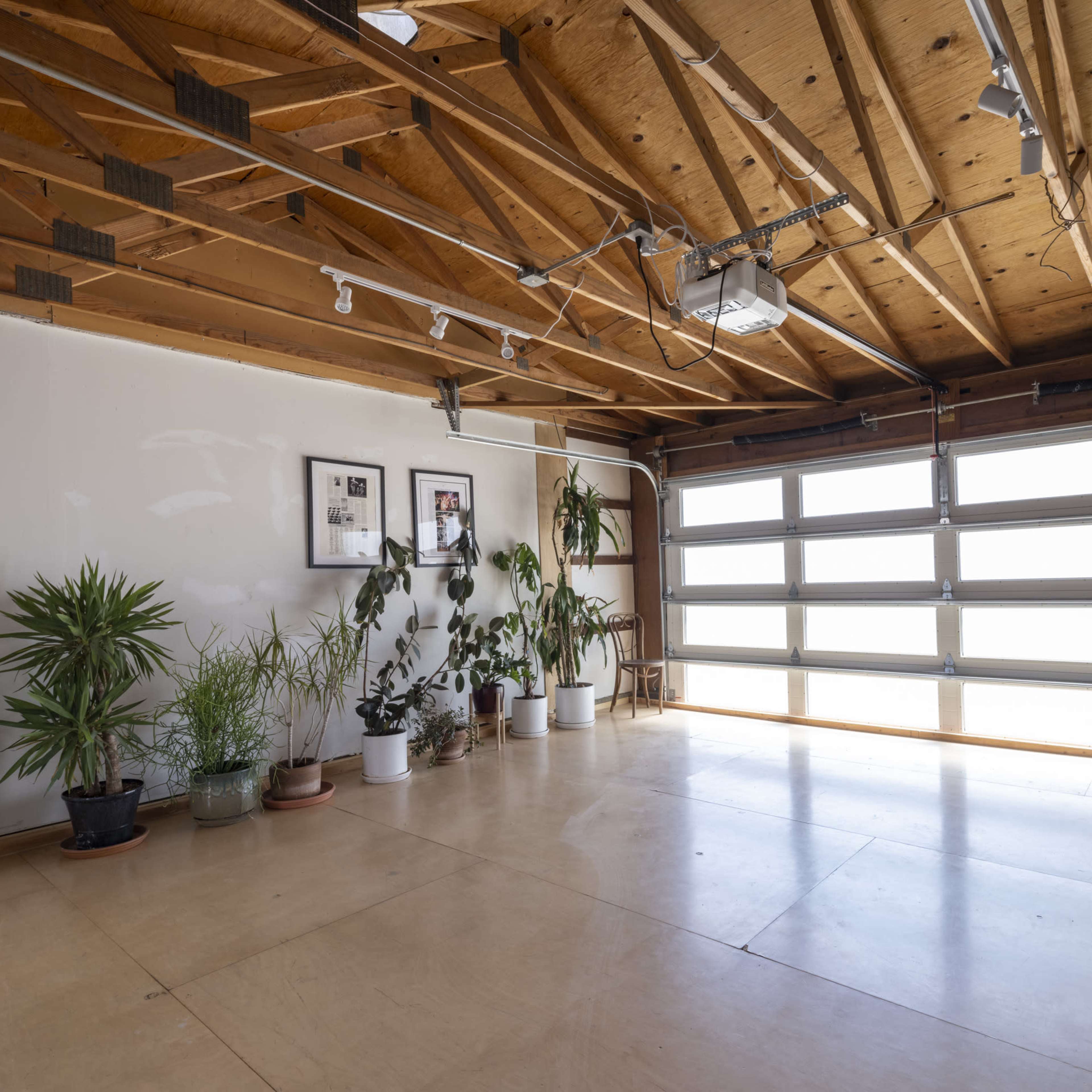 A bright, empty garage with wooden beams on the ceiling, large windows, and several potted plants along one wall.