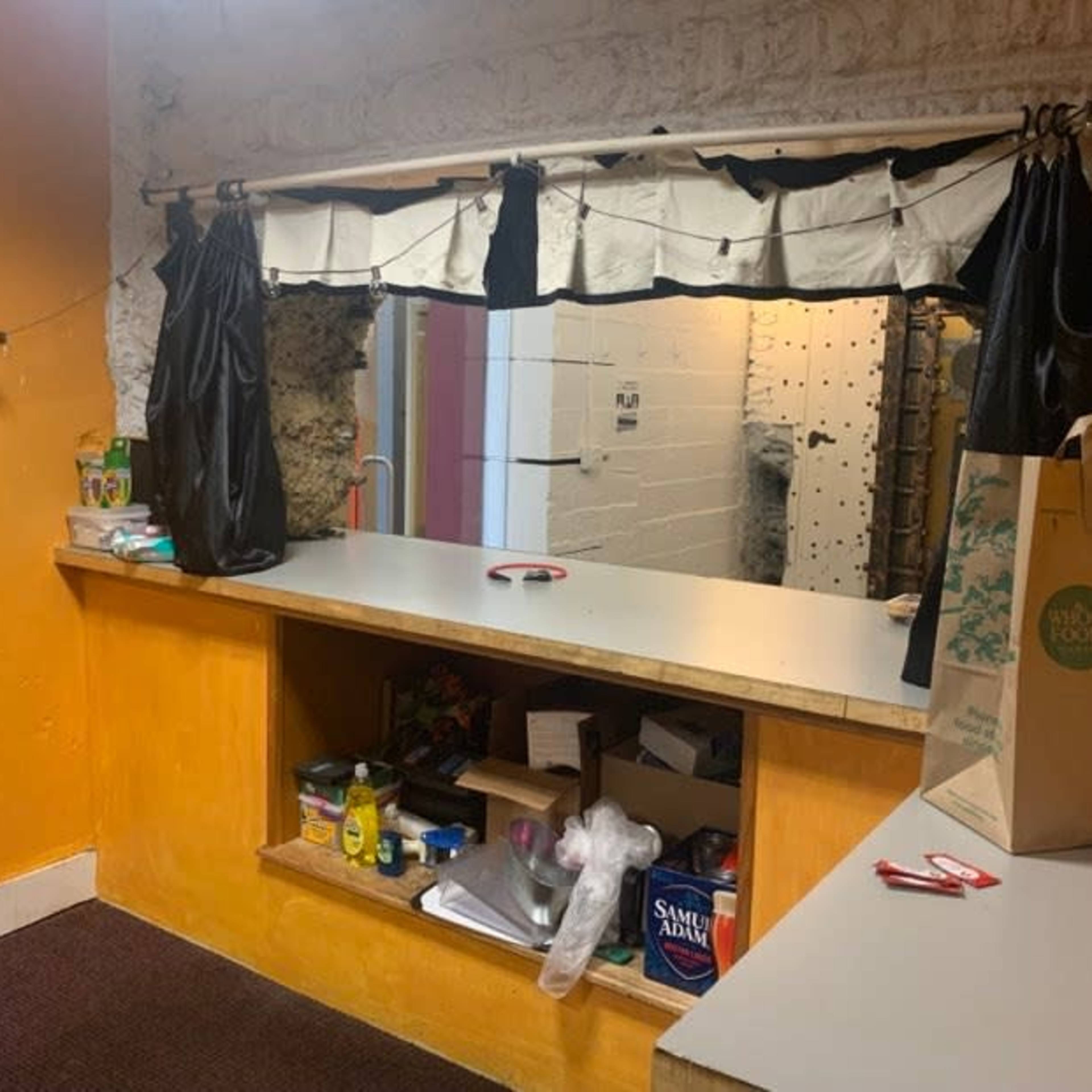 A small room with an orange wall, a counter with various items, and a window covered by black curtains.