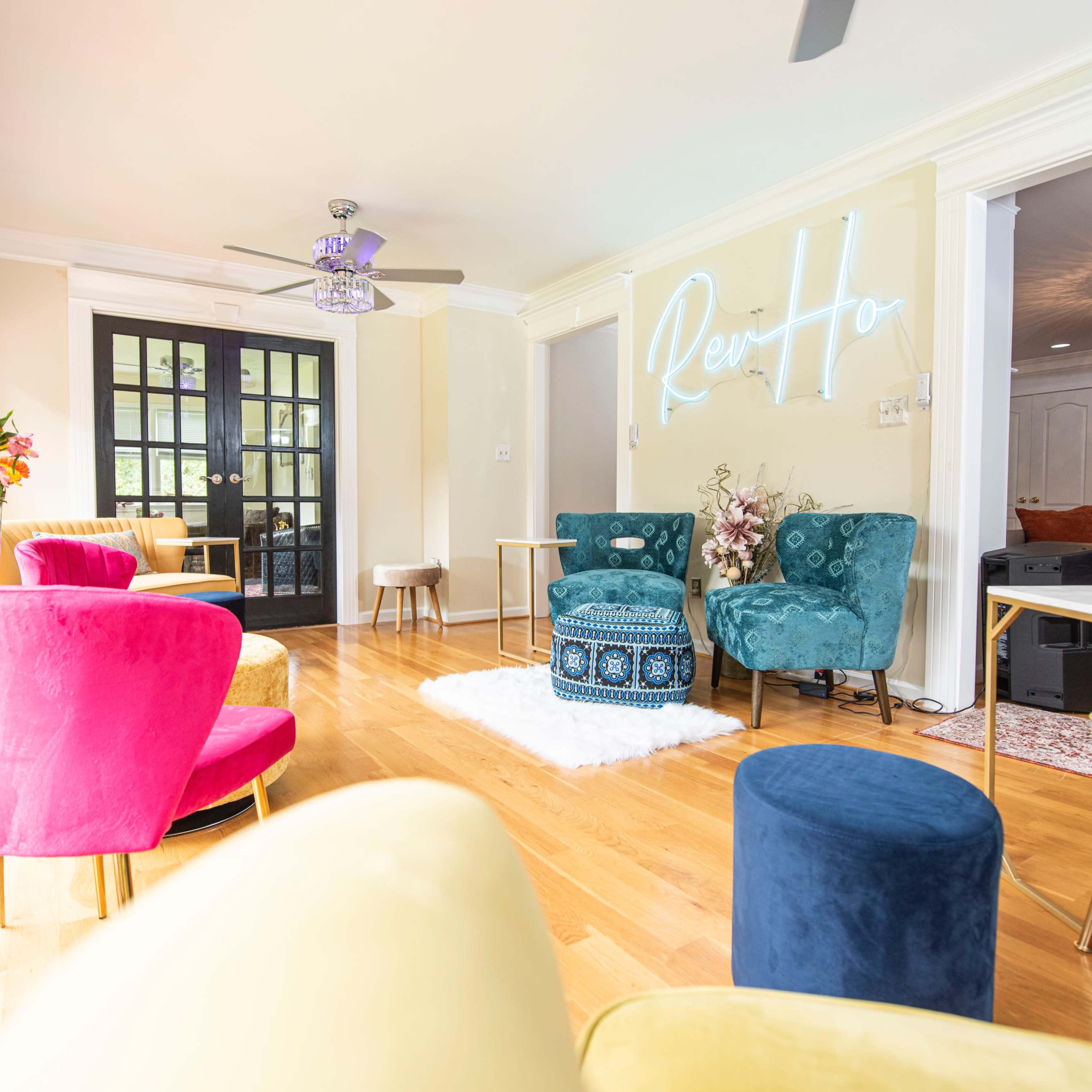 The bright room features colorful chairs, a small table, and a floral arrangement beside a wall with a neon sign.