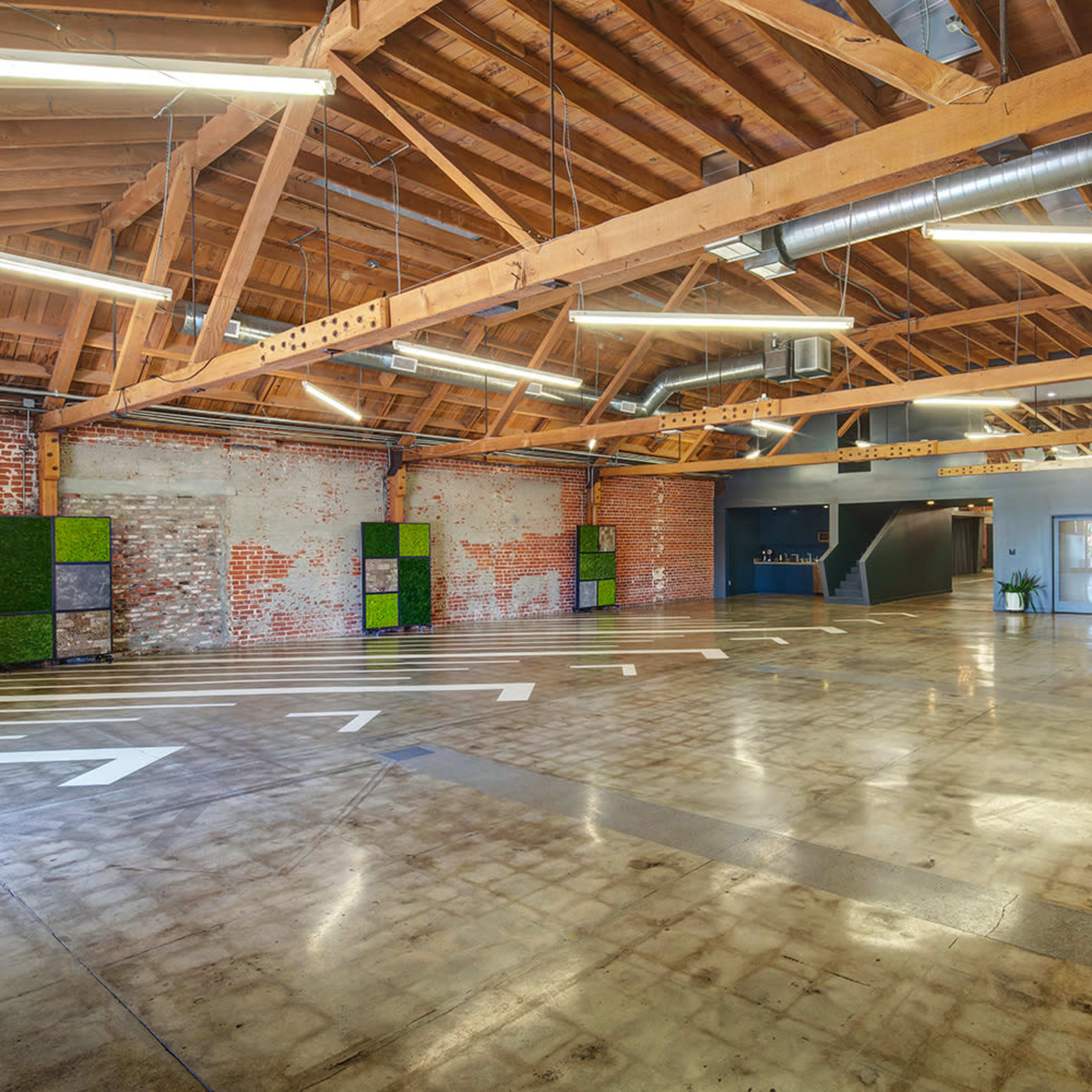 A spacious industrial-style room with exposed wooden beams, polished concrete flooring, and brick walls, featuring several decorative plants along the edges.
