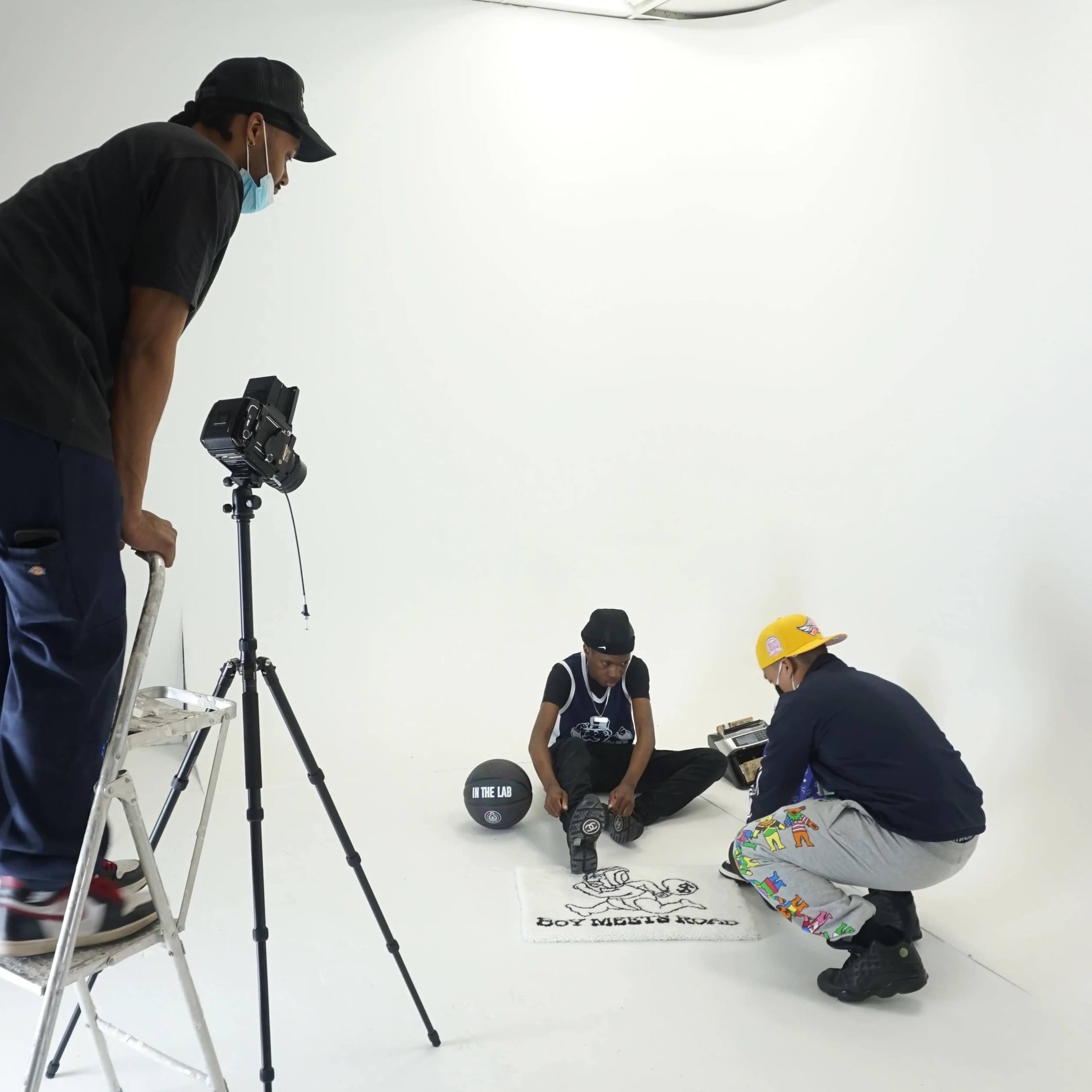 A person on a ladder sets up a camera while two individuals prepare for a photoshoot on a white background.