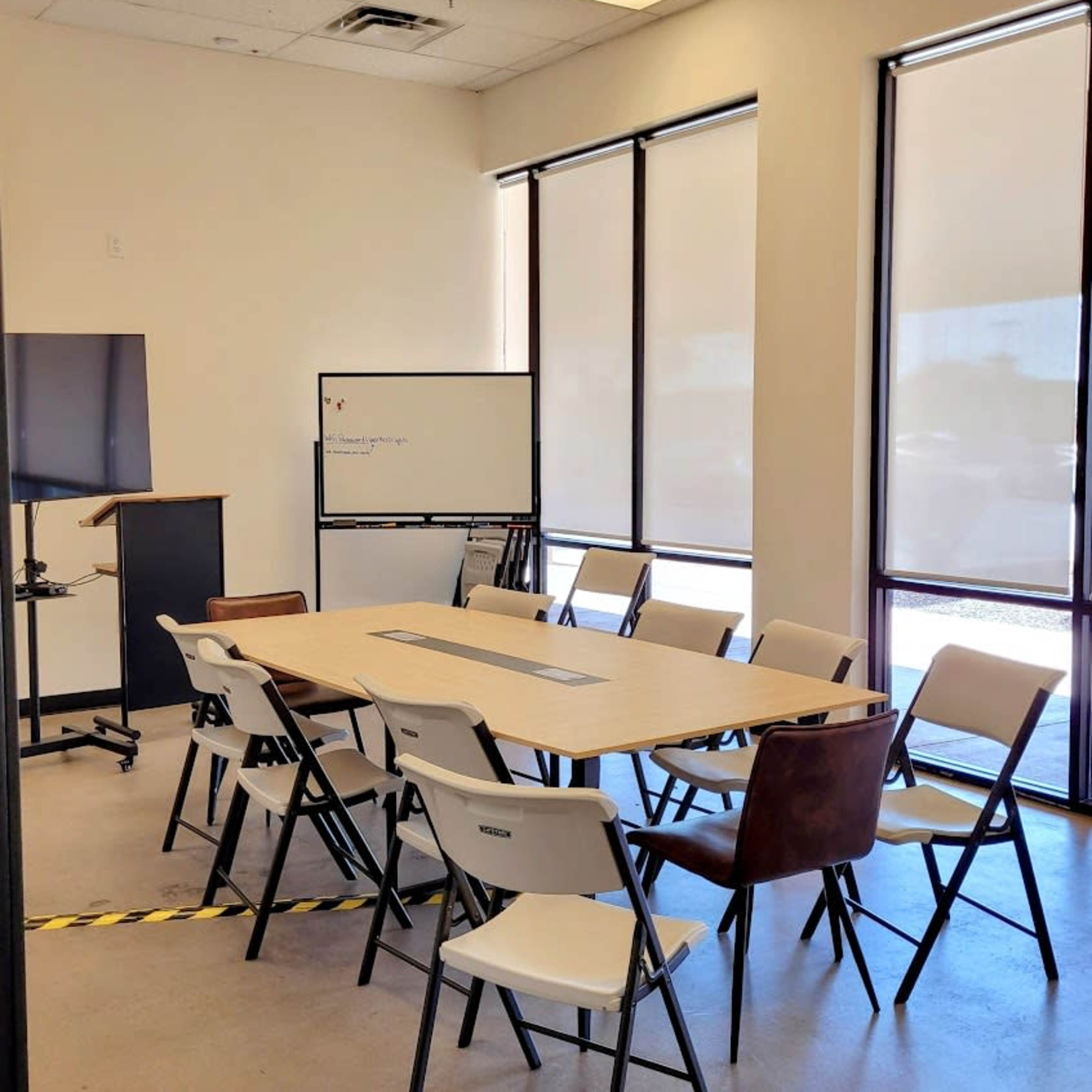 A conference room features a large table surrounded by chairs, a whiteboard, and a television on a stand, with natural light filtering through large windows.