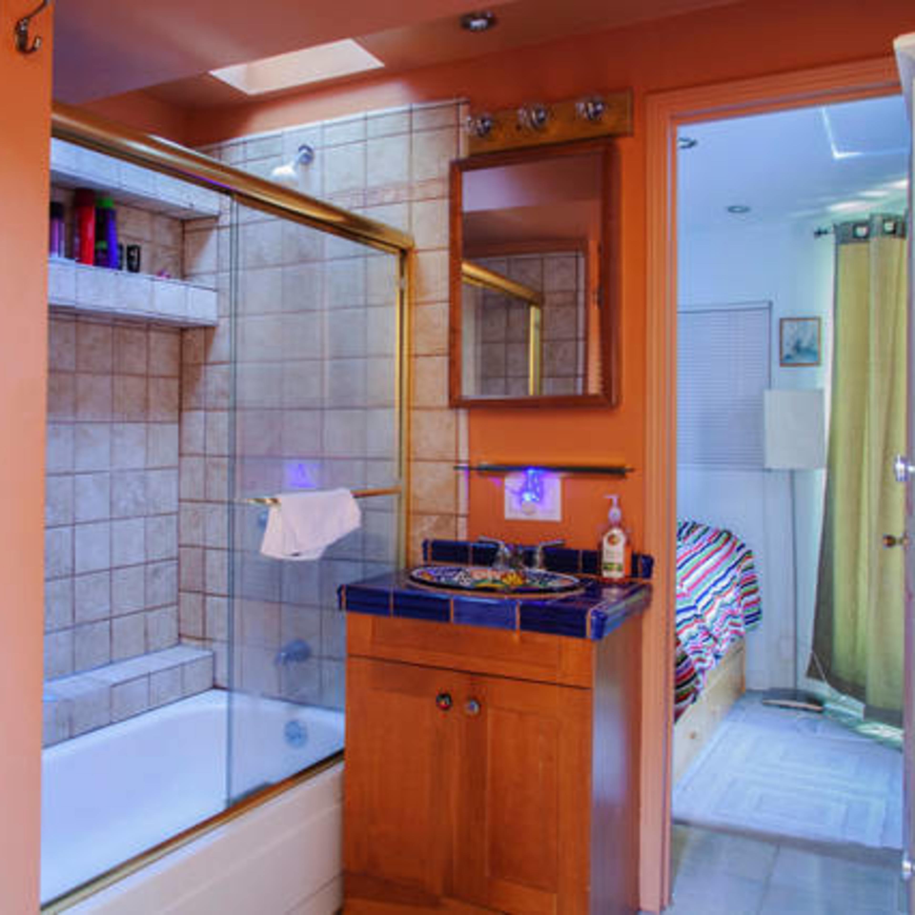 A bathroom with a shower area featuring a glass door, a vanity with a sink, and a nearby room containing a bed and a window with curtains.