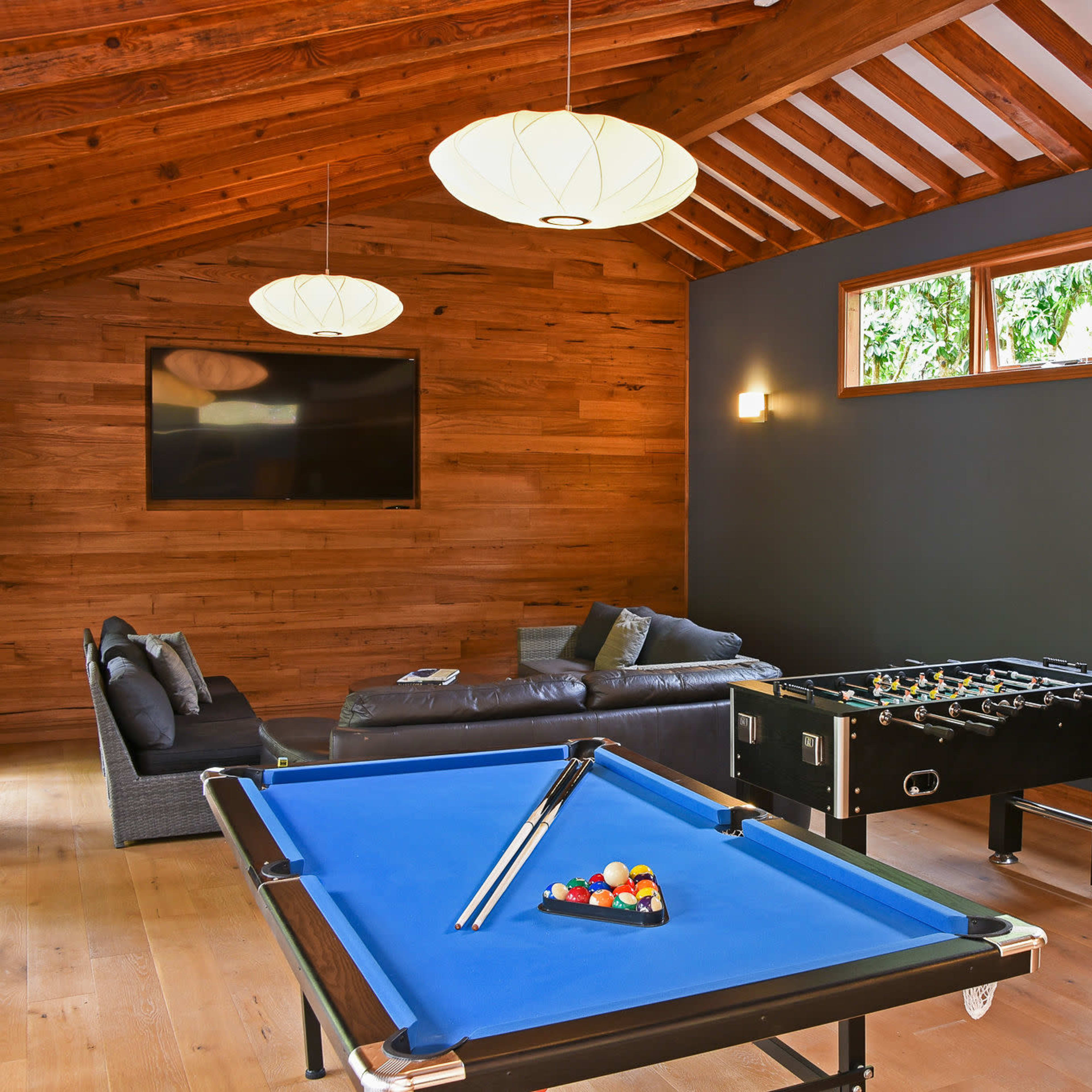 A game room features a blue pool table, a foosball table, and a sectional sofa under wooden beams with large windows allowing natural light.
