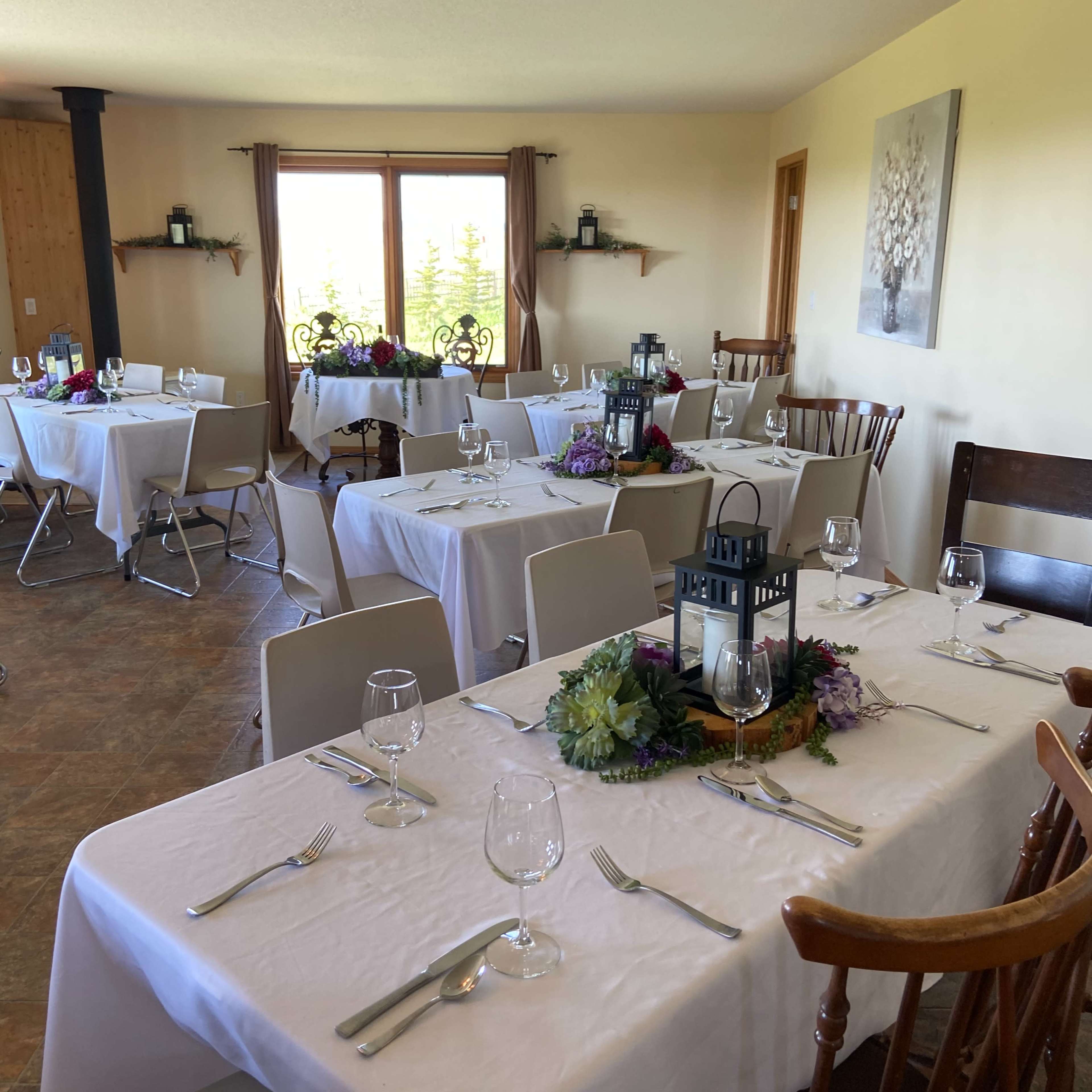 A dining room with several tables set with white linens, glassware, and floral decorations, ready for a gathering or event.