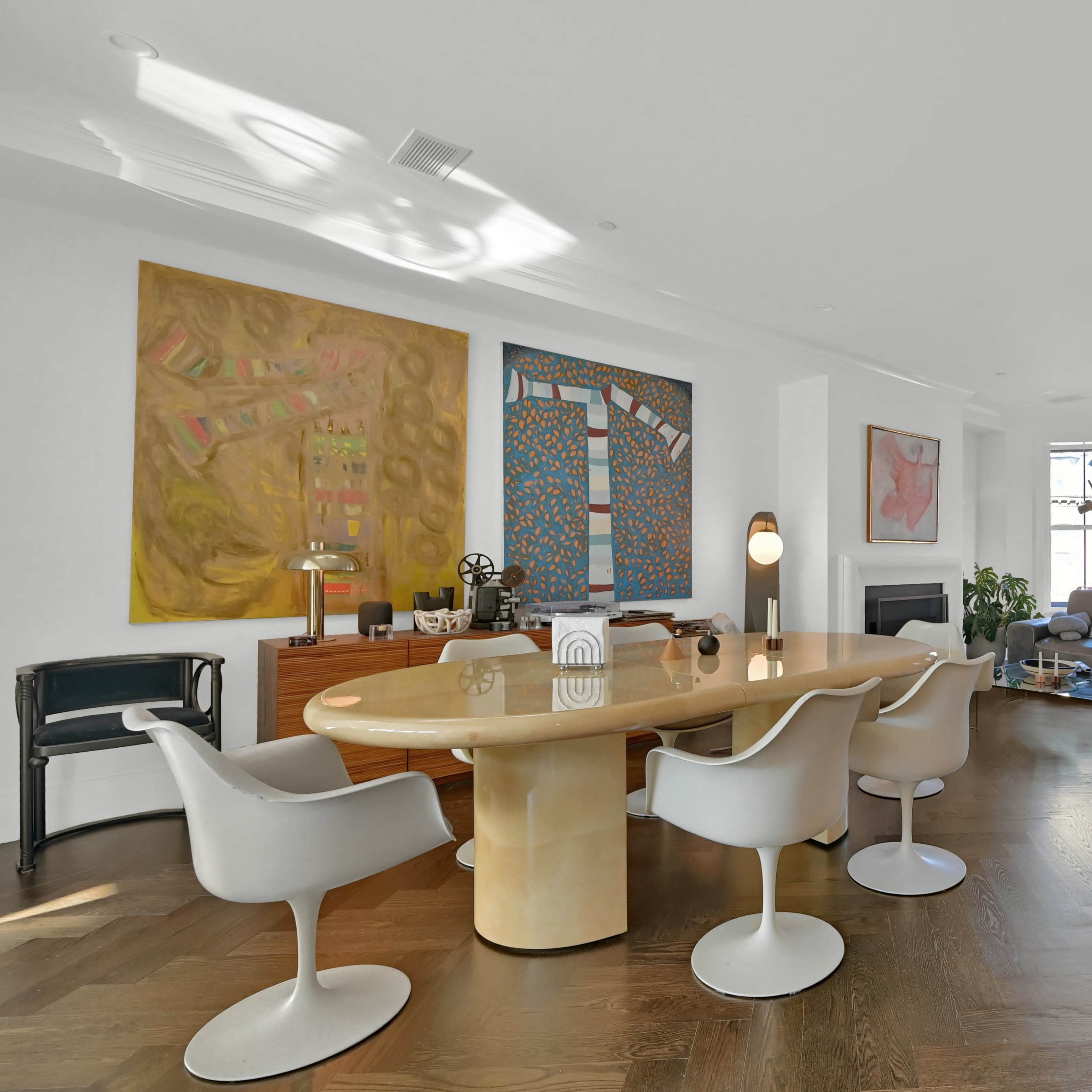 A modern living room with a round dining table surrounded by white chairs, two large paintings on the walls, and a gray sofa facing a fireplace and windows.