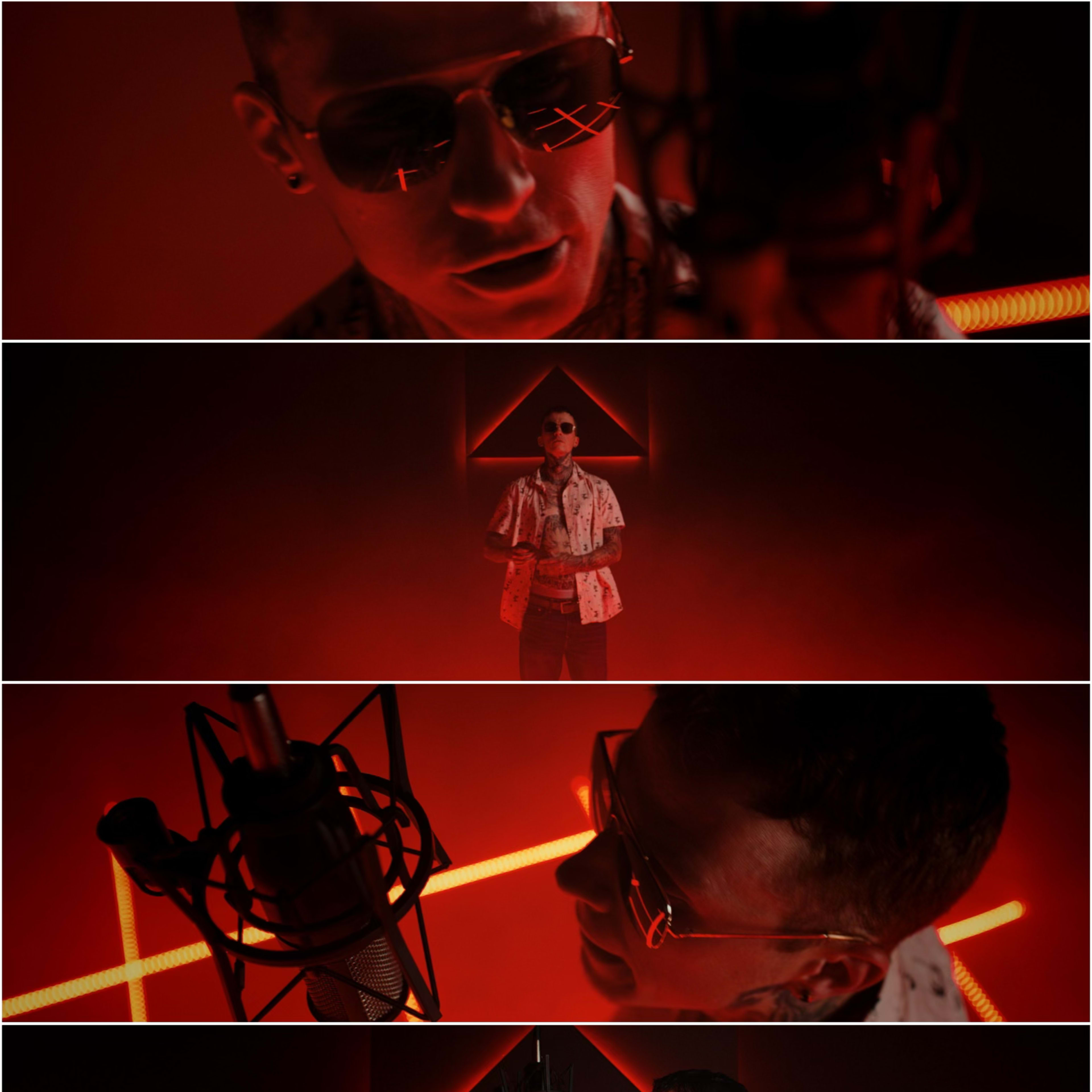 A person wearing sunglasses stands in front of a microphone, illuminated by red lights and geometric shapes in a dimly lit environment.