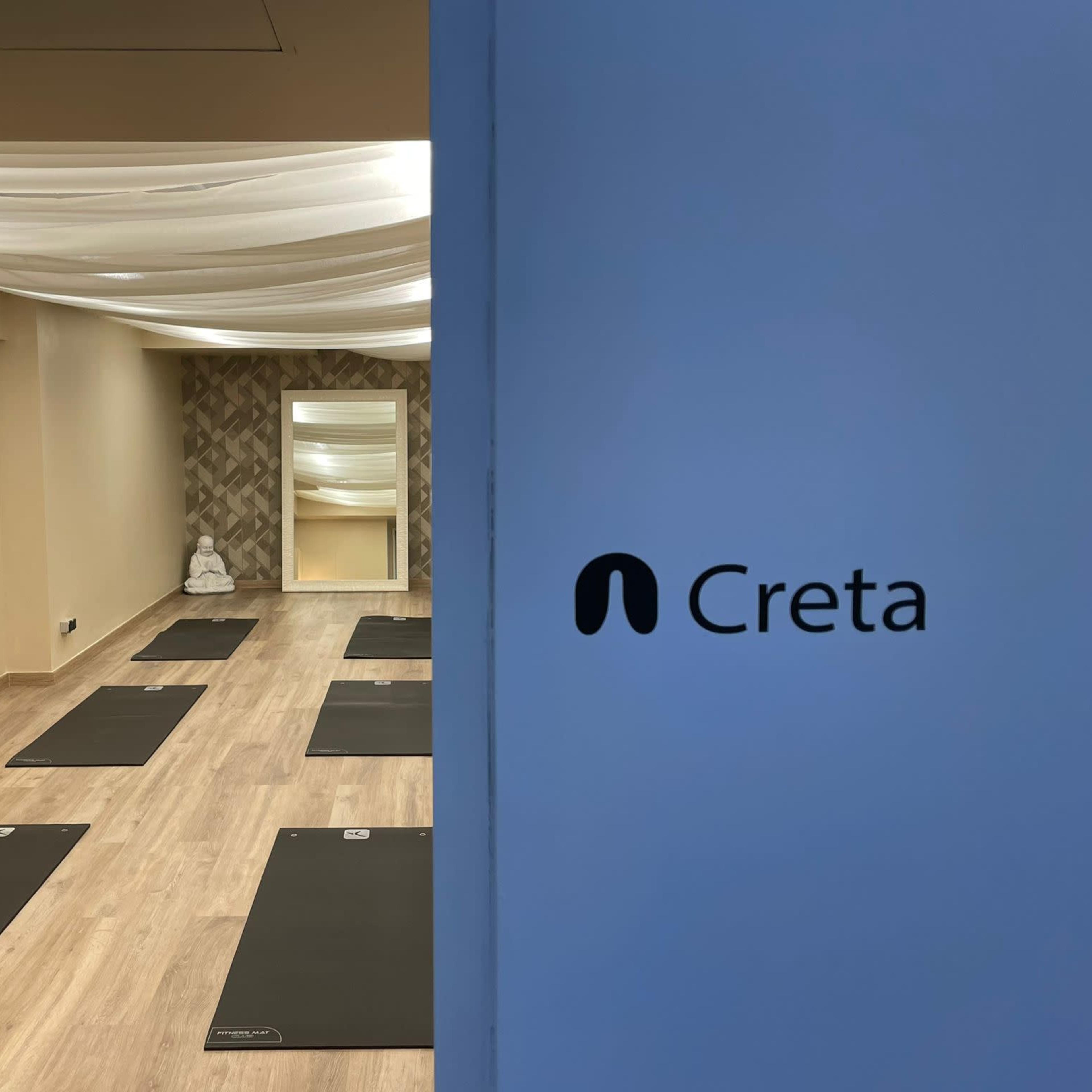A yoga studio with several exercise mats arranged on the wooden floor and a large mirror on the wall, with a sign reading "Creta" visible.