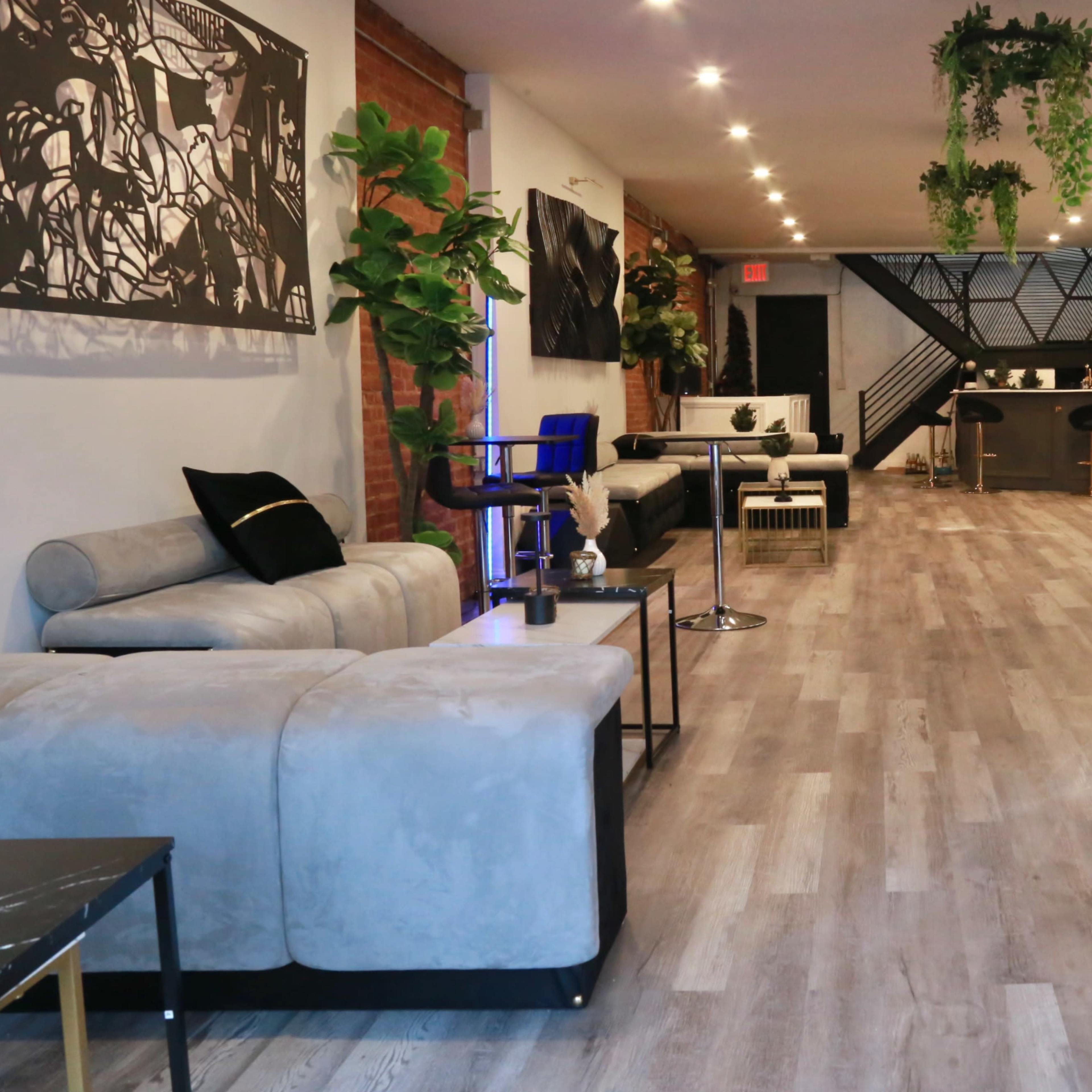 A modern, spacious lounge area with comfortable seating, wooden flooring, and plants, illuminated by overhead lights.