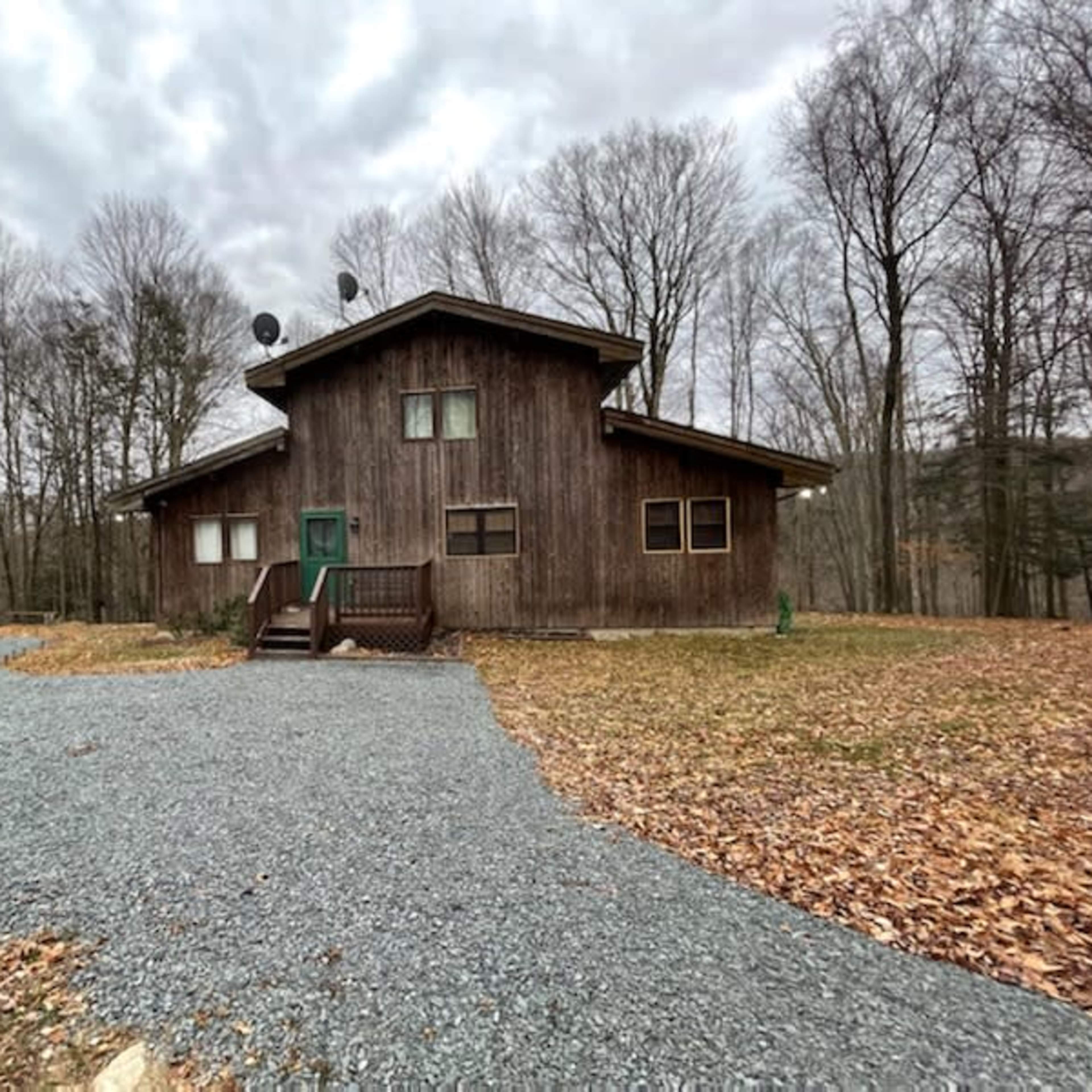 A wooden house with a sloped roof and green door is situated in a gravel-lined clearing surrounded by trees.