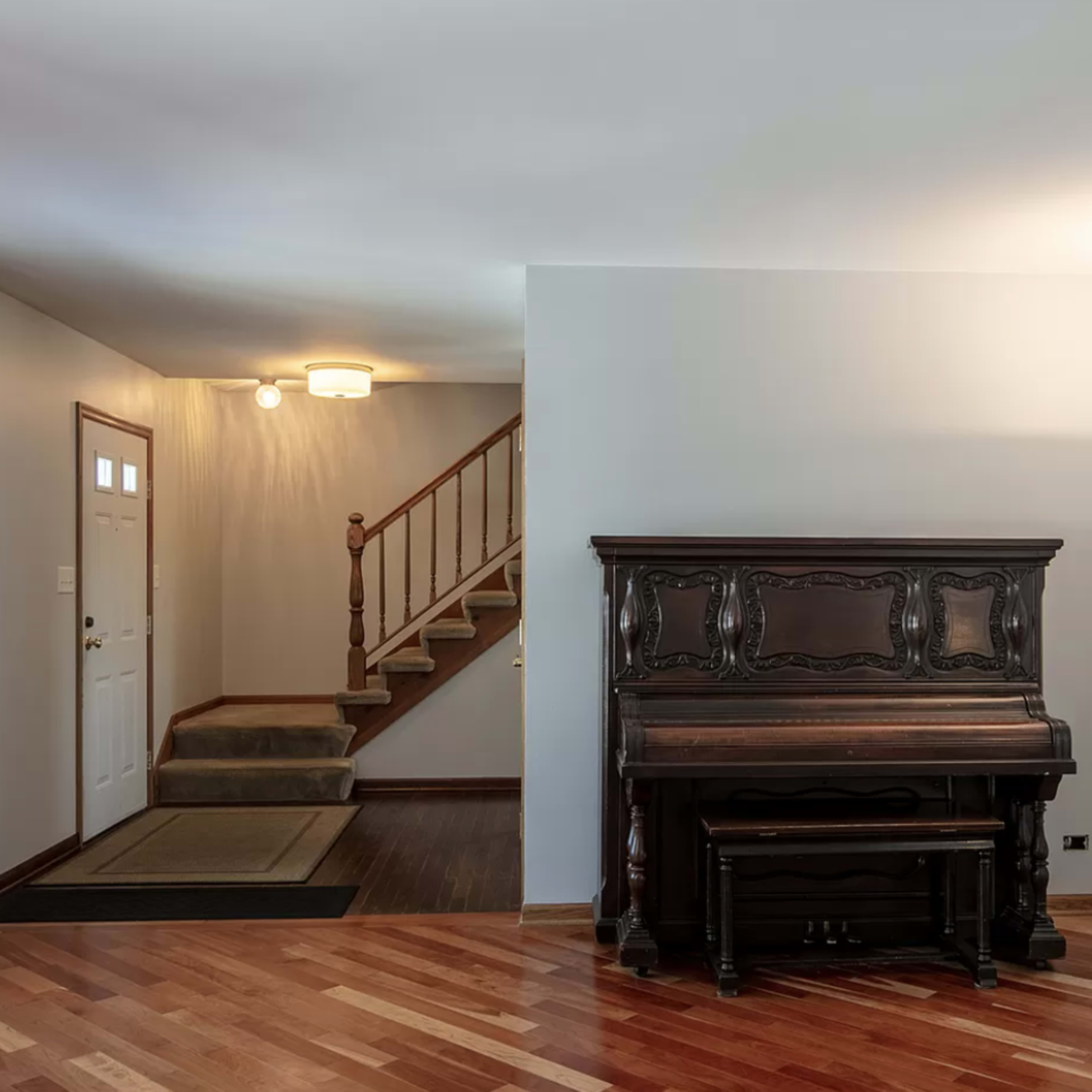 A living space featuring a wooden piano, a staircase, and a front door leading to an entryway.