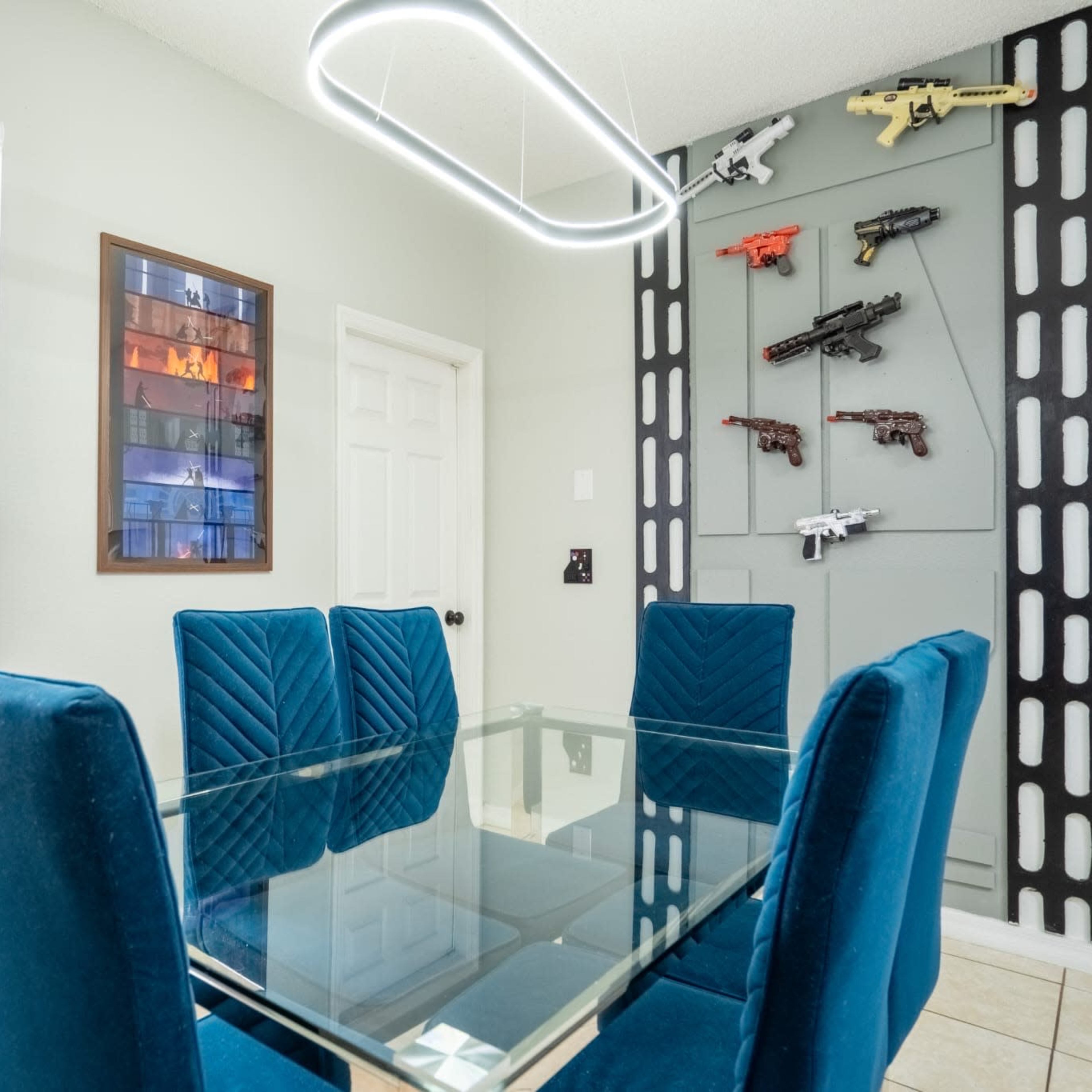 A modern dining area features a glass table surrounded by blue upholstered chairs, with a wall displaying replica firearms and a framed artwork.