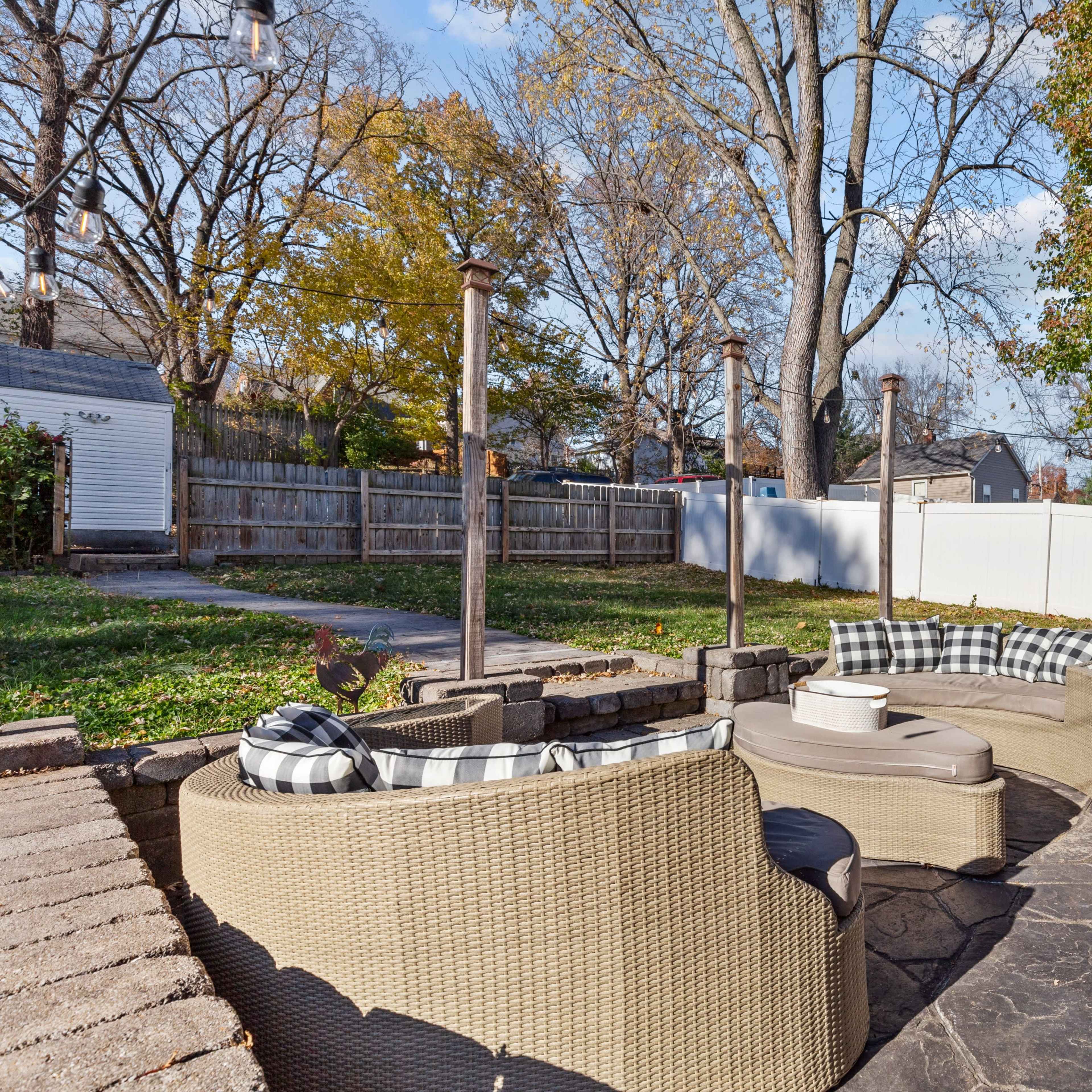 A backyard patio with stone flooring, featuring a seating area with wicker chairs and a round table, surrounded by grass and trees.