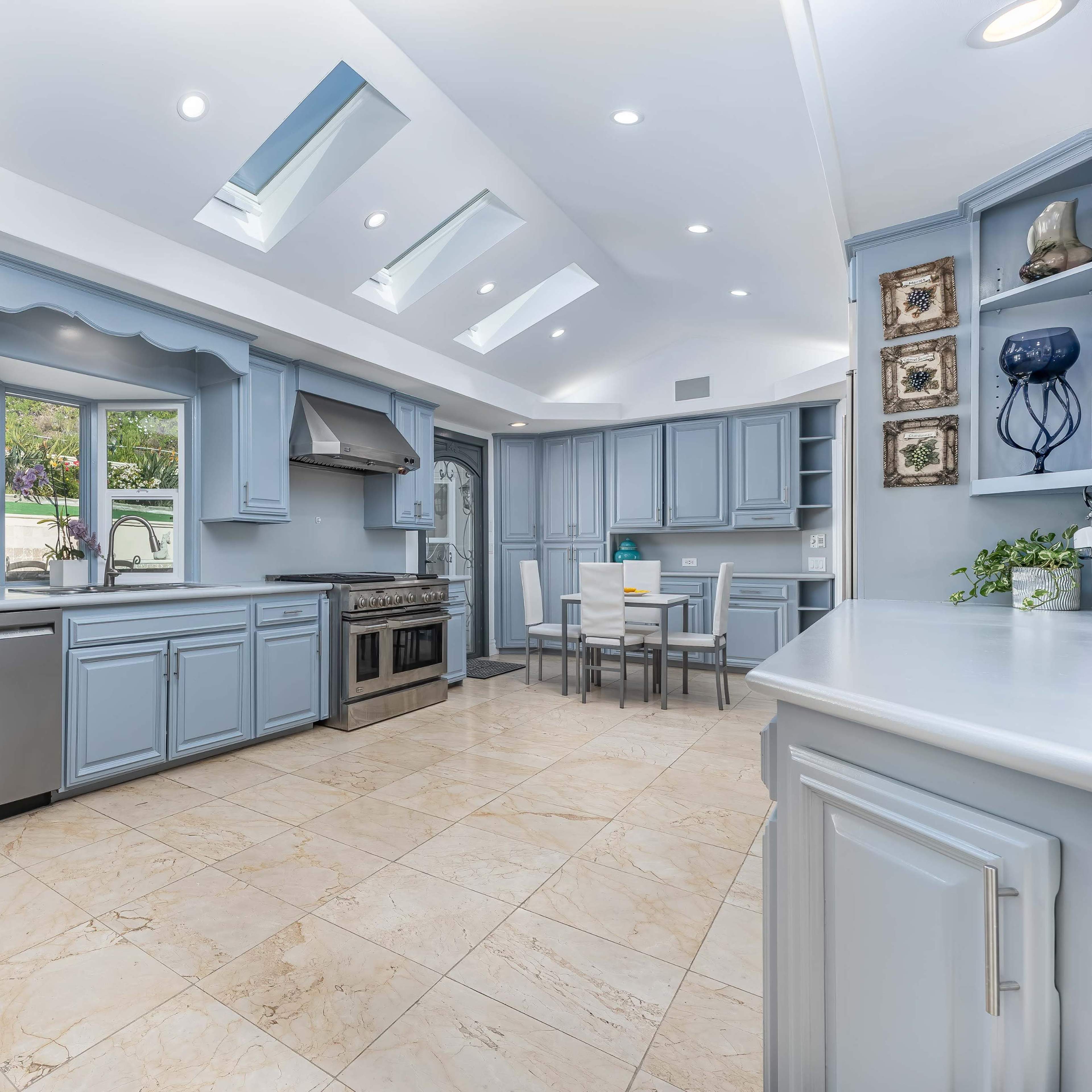 A spacious kitchen features blue cabinets, stainless steel appliances, and skylights illuminating the marble-tiled floor.