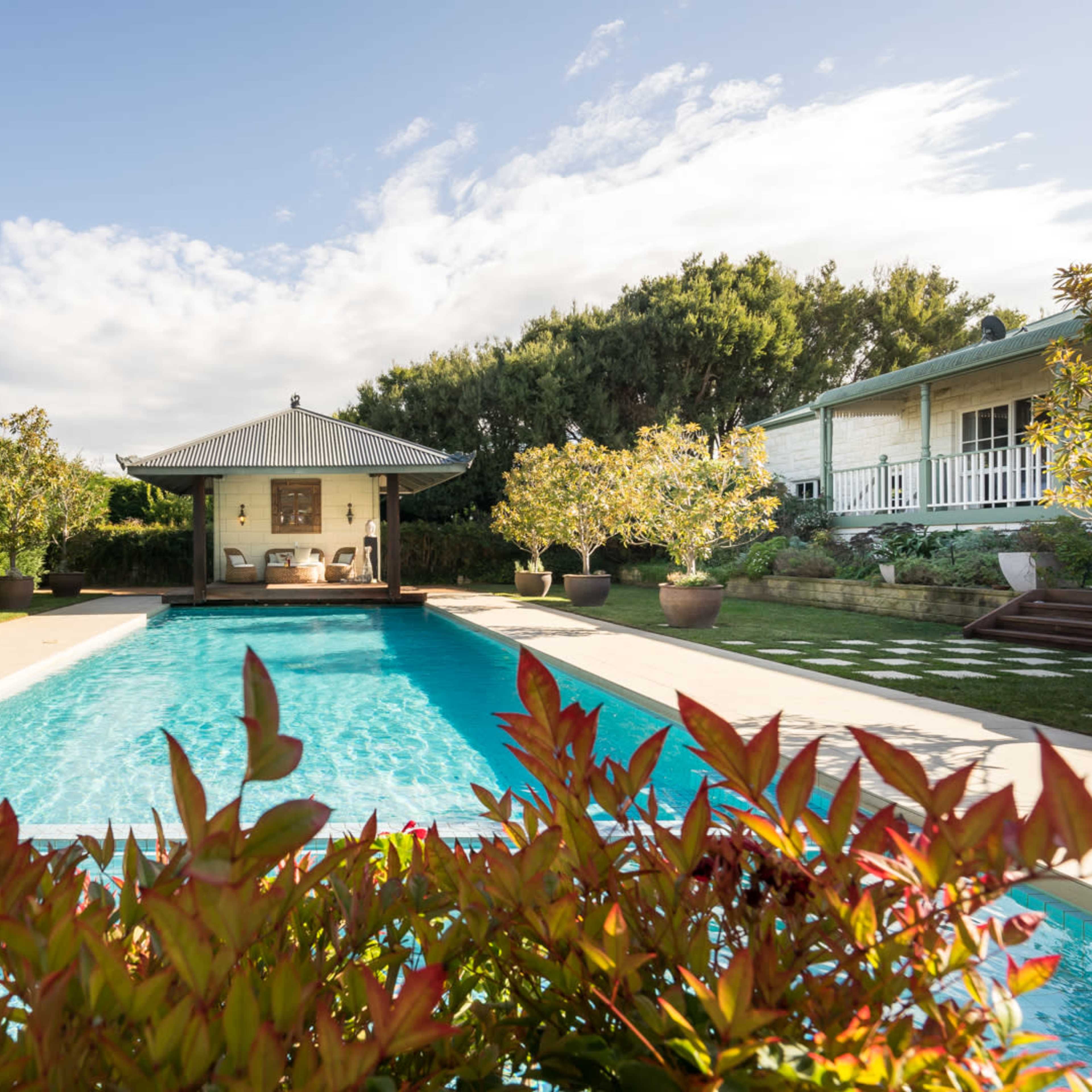 A rectangular swimming pool is surrounded by lush greenery and decorative plants, with a gazebo and house visible in the background.