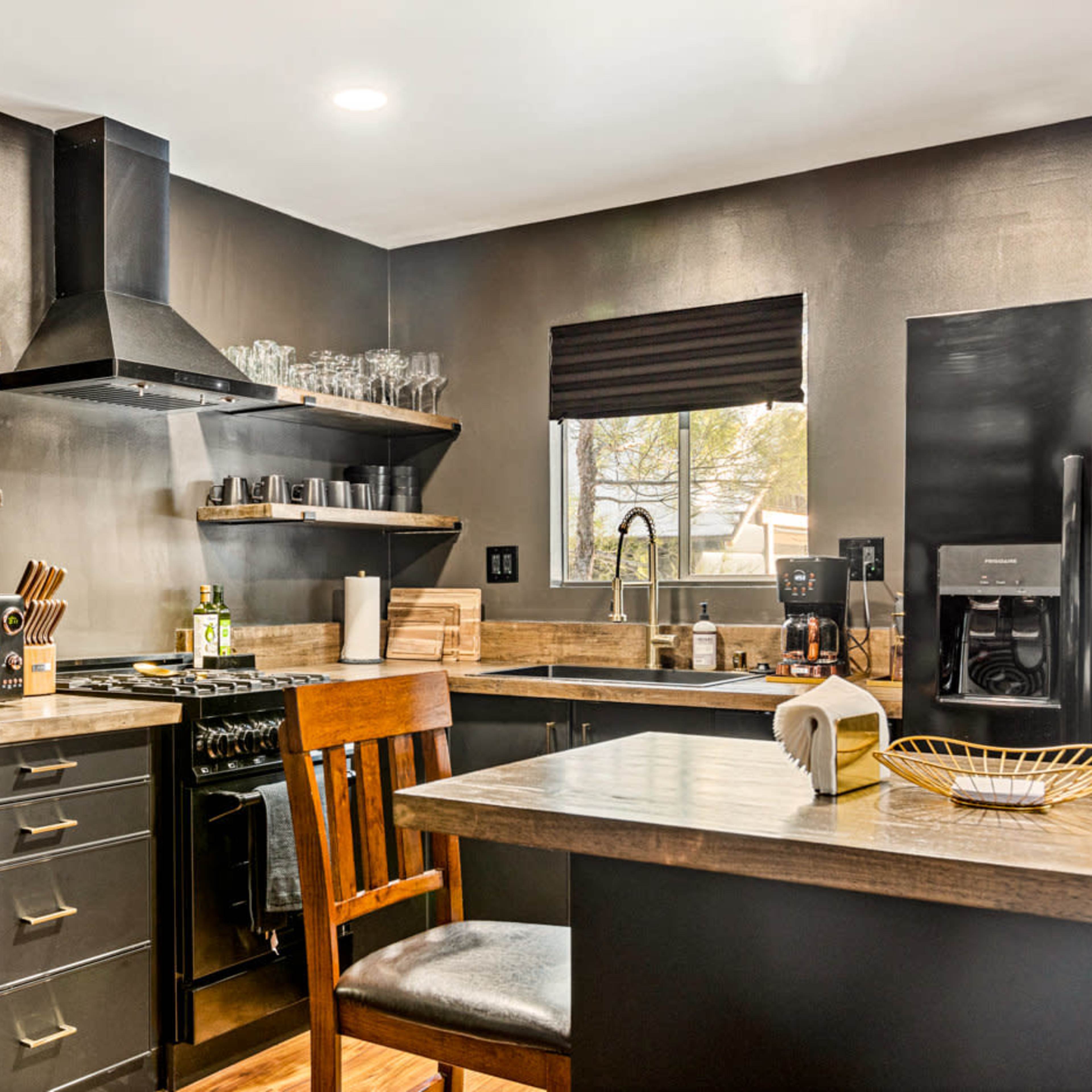 A modern kitchen features dark cabinetry, wooden shelves, and stainless steel appliances, with a dining chair positioned at a countertop.