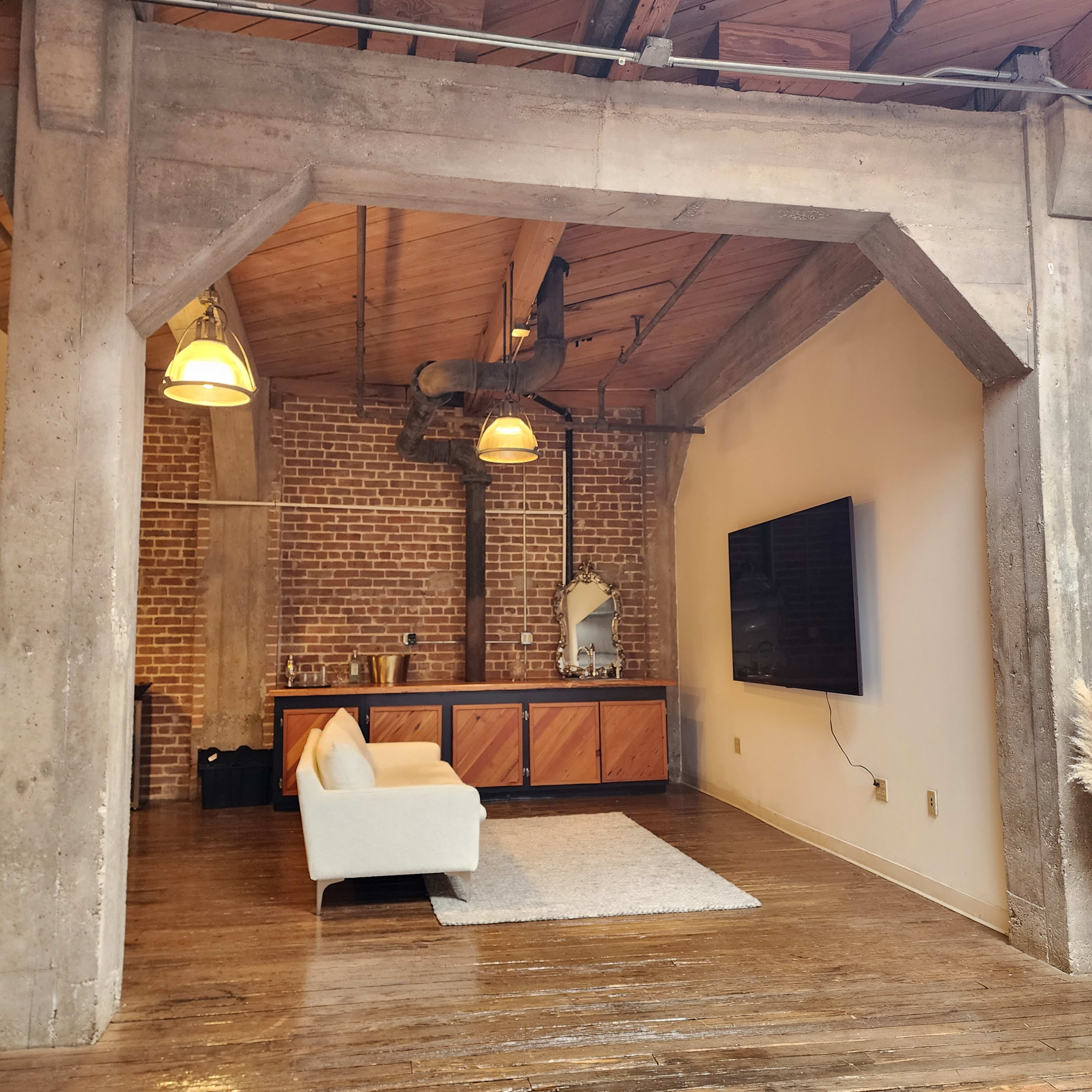 A spacious living area with exposed brick walls, a wooden ceiling, a modern sofa, a flat-screen TV mounted on the wall, and a wooden cabinet.