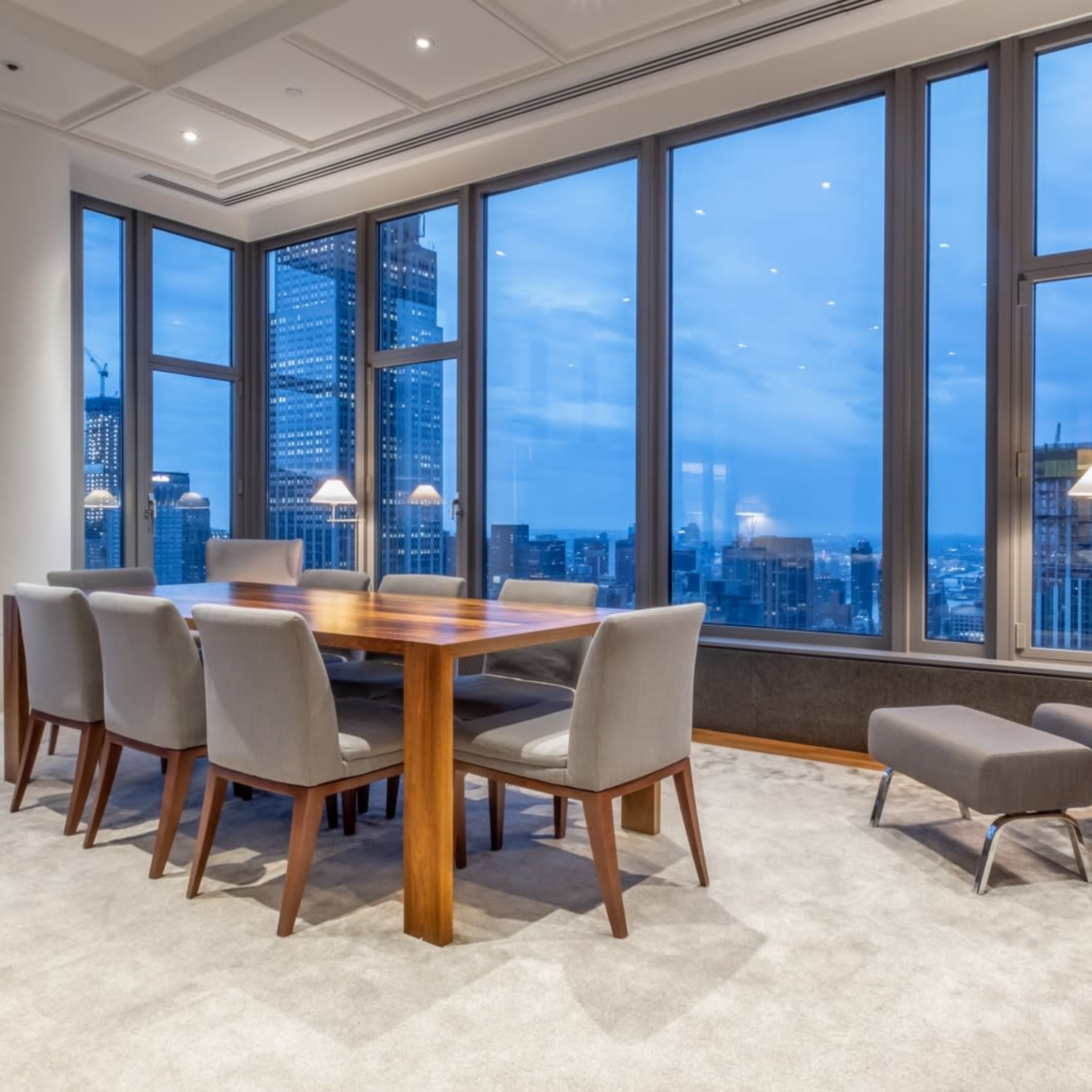 A modern conference room features a wooden table surrounded by gray chairs, with large windows showcasing a city skyline at twilight.