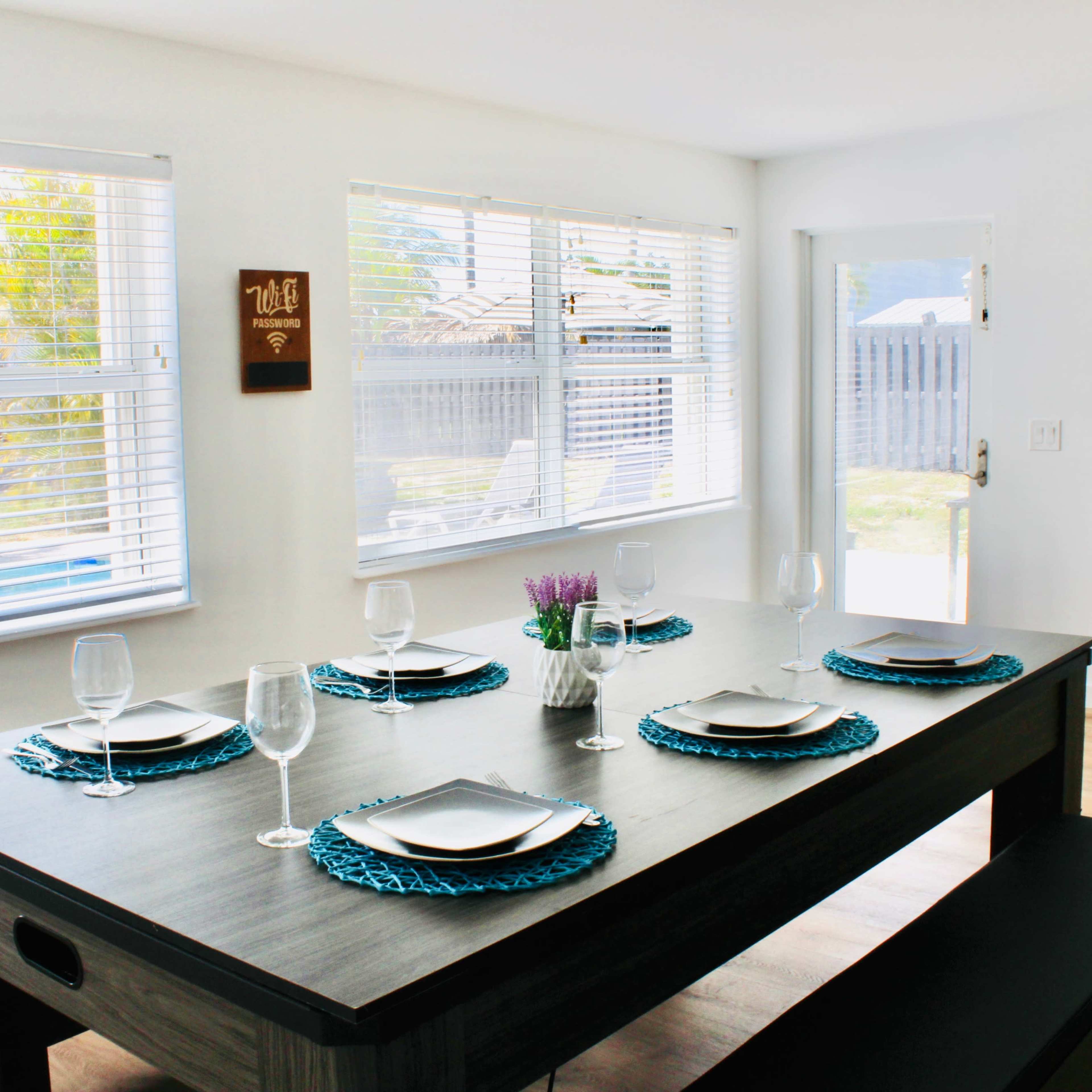 A modern dining room features a rectangular table set with plates, glasses, and decorative placemats, surrounded by large windows that provide ample natural light.