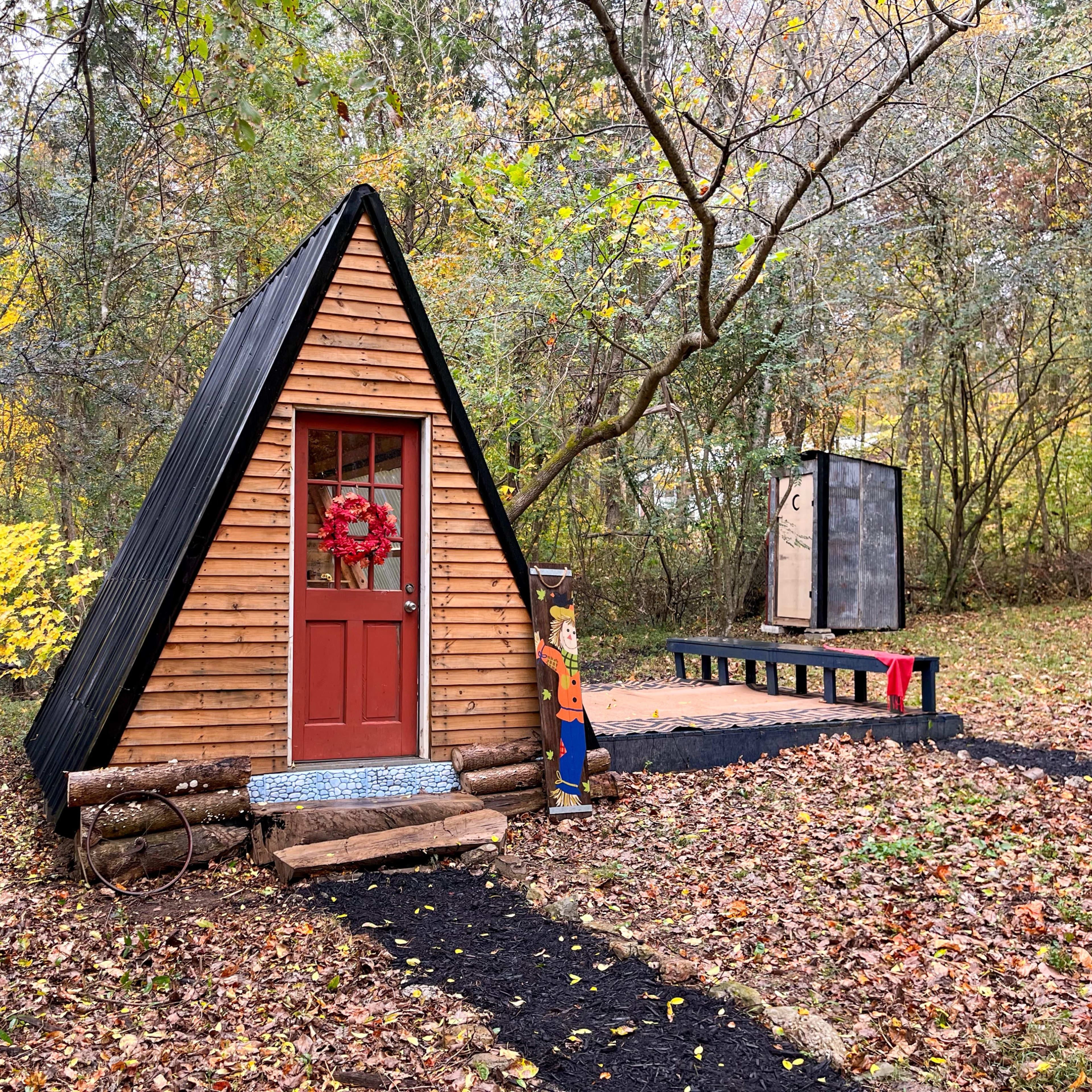 A small A-frame cabin with a red door and window, surrounded by autumn foliage and a path leading to a platform.