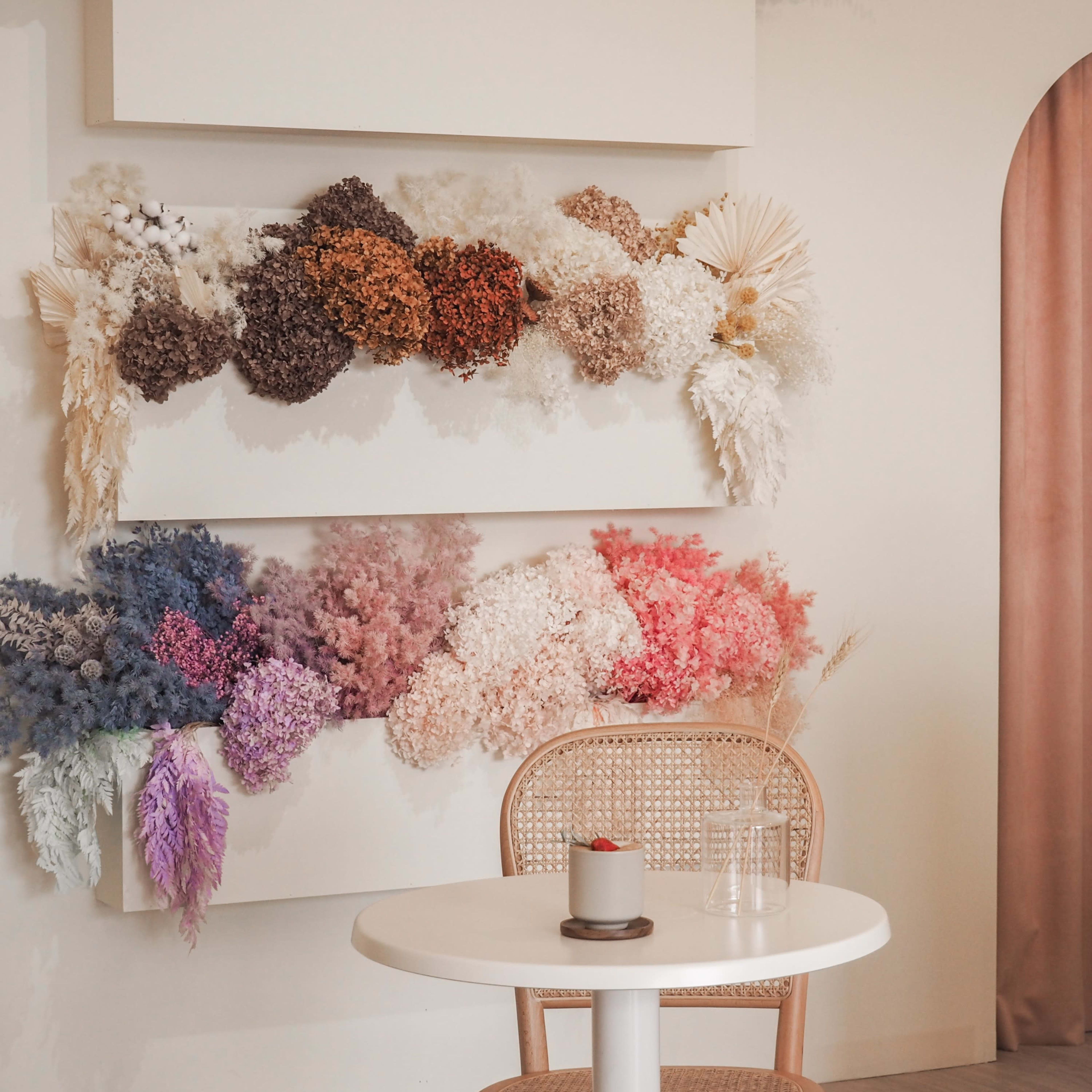 A minimalist interior features a white wall adorned with a variety of dried flowers, a round table with a small dessert, and a beige curtain in the background.