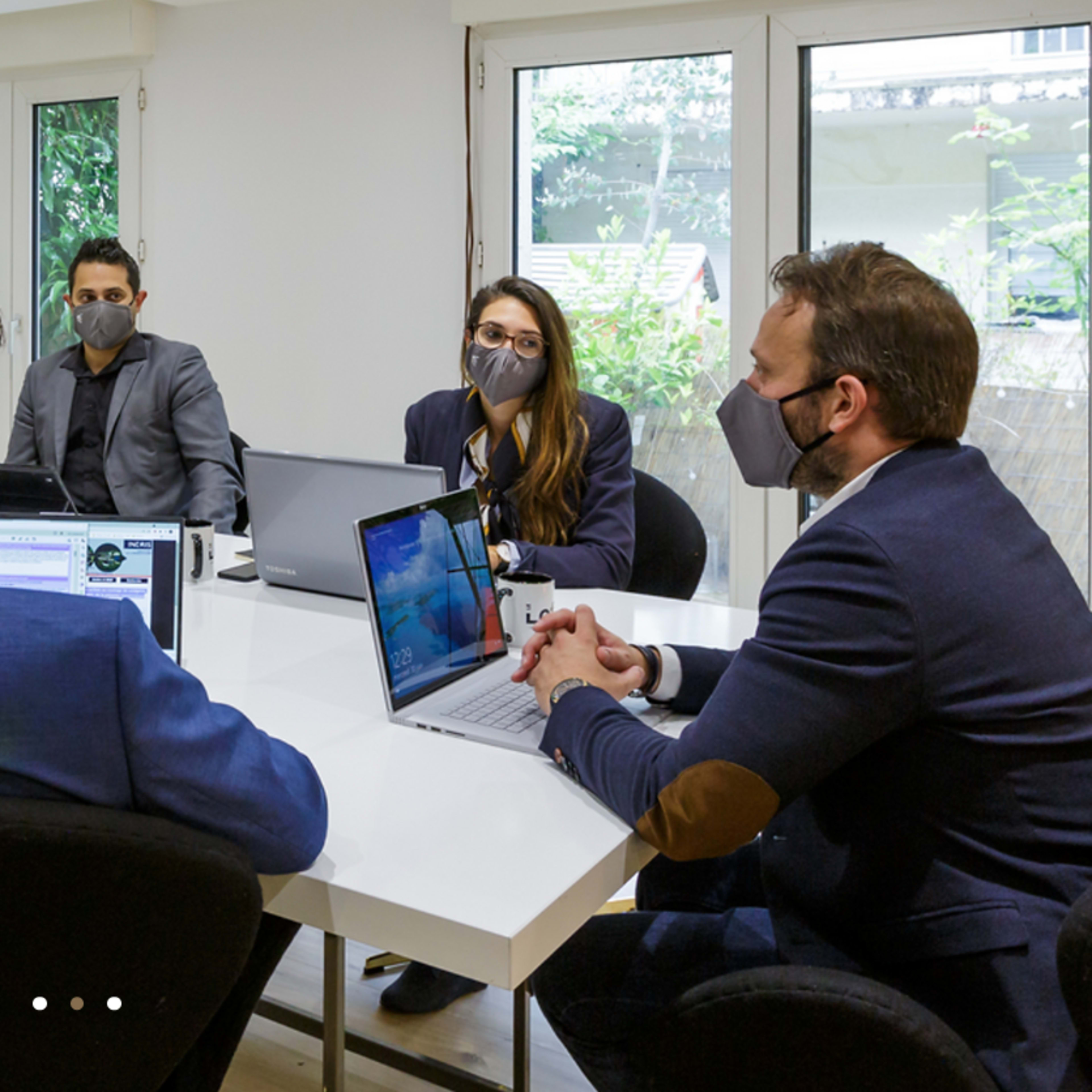 A group of six people wearing masks is seated around a conference table with laptops and documents in a well-lit meeting room.