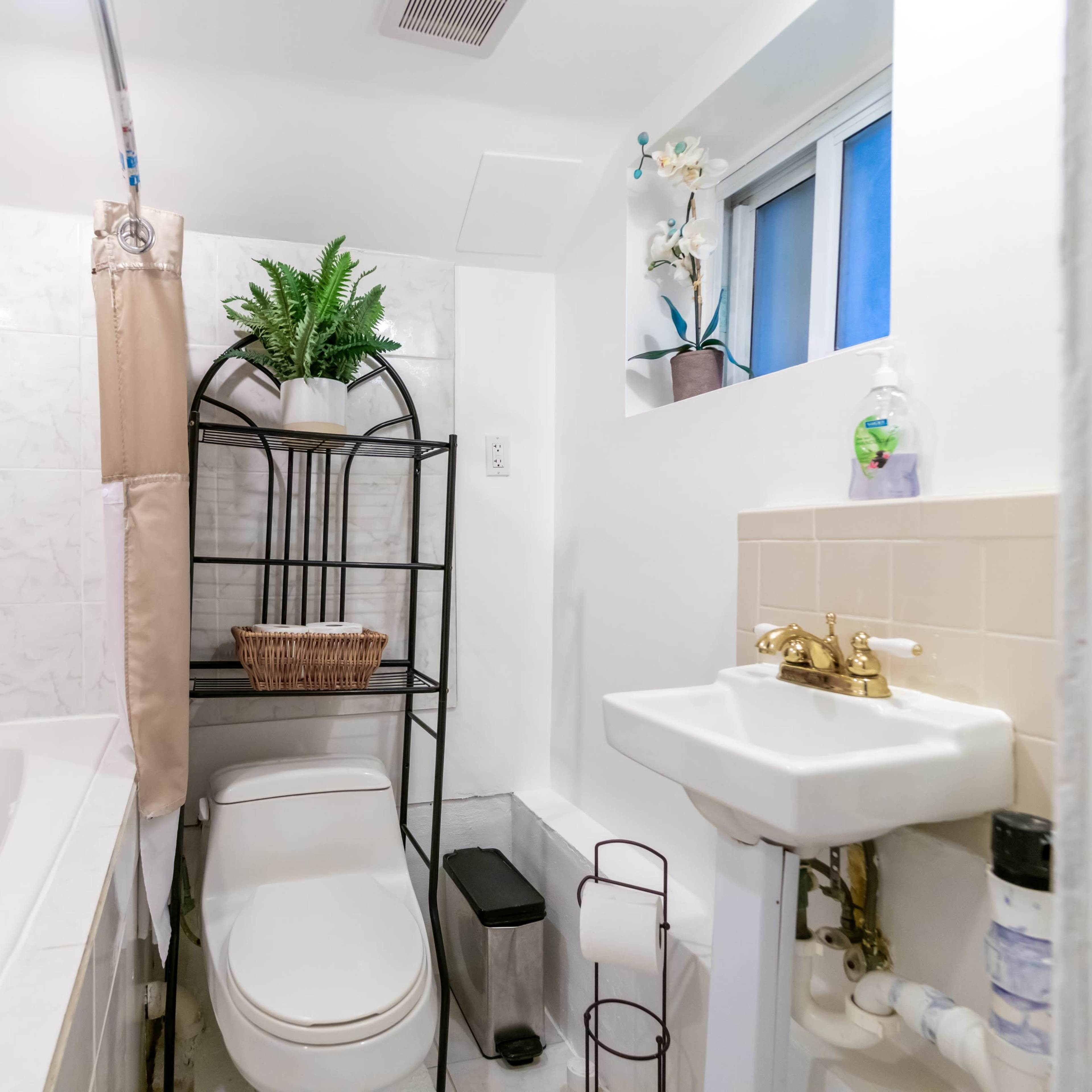 A small bathroom featuring a bathtub, a toilet, a sink, and a storage rack with potted plants.