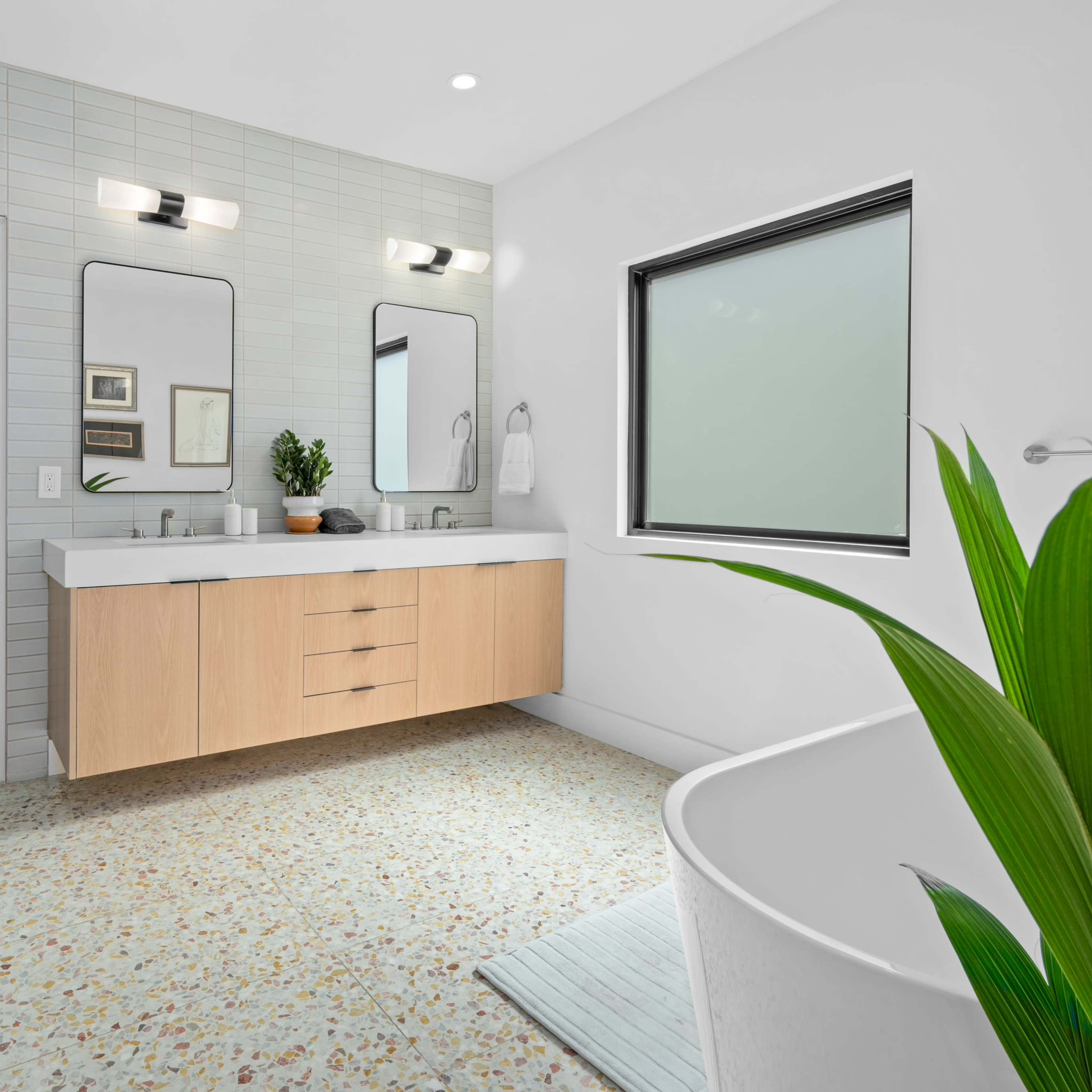 A modern bathroom features a double vanity with wooden cabinetry, two mirrors, a freestanding bathtub, and a potted plant by the window.