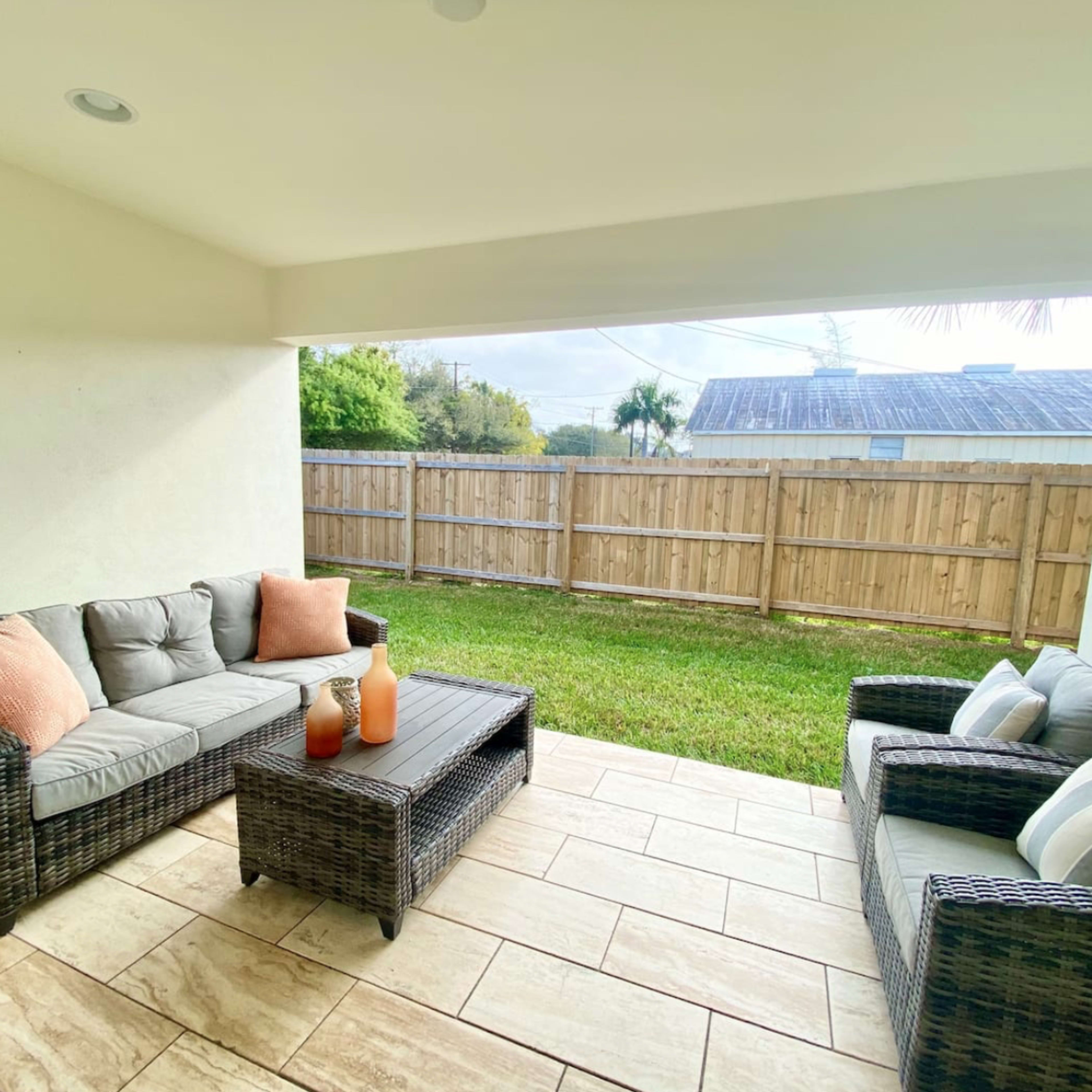 A covered patio features a seating area with wicker furniture and a coffee table, overlooking a grassy yard enclosed by a wooden fence.